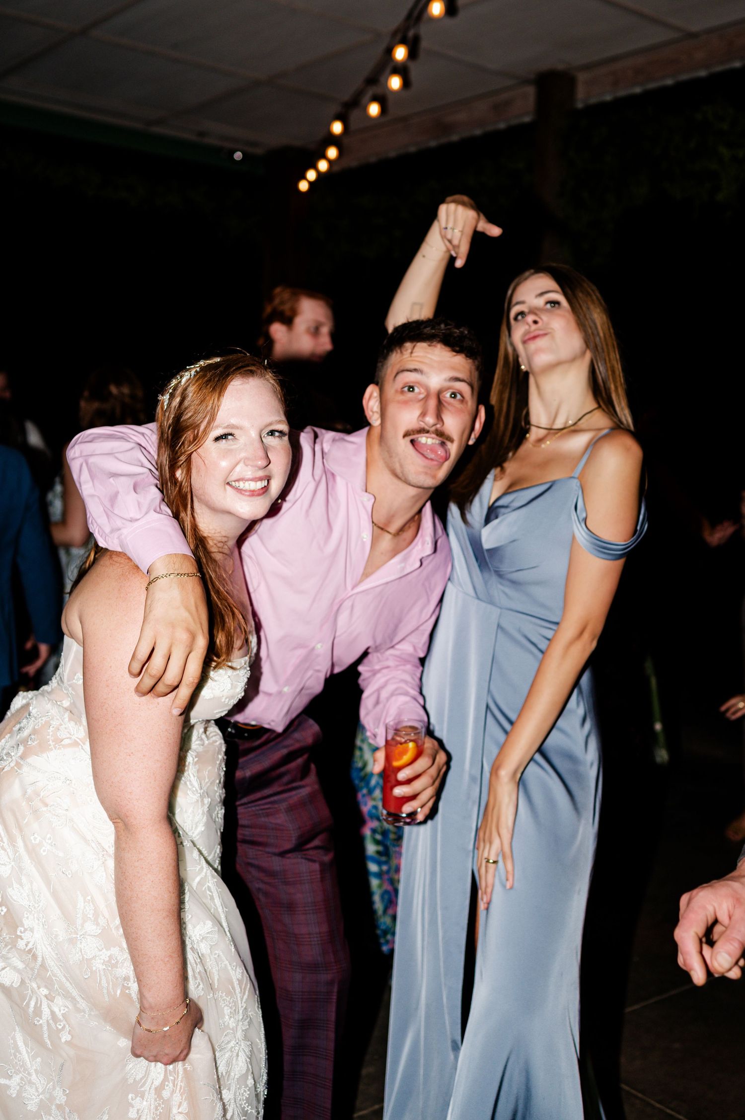 A bride in a lace gown laughs with two guests while dancing at a lively wedding reception.