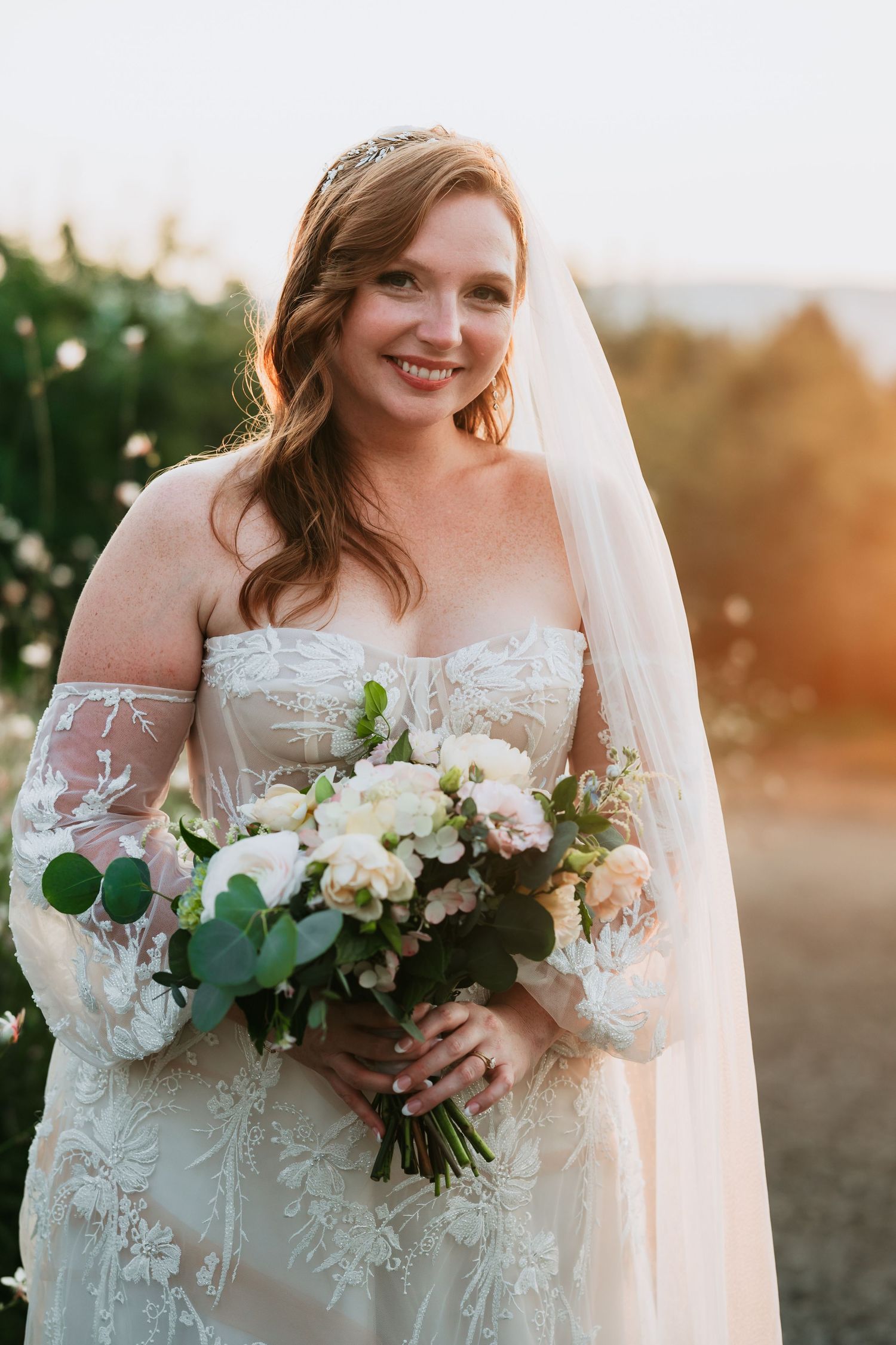 Smiling bride in lace gown holds white bouquet at golden hour in outdoor setting.