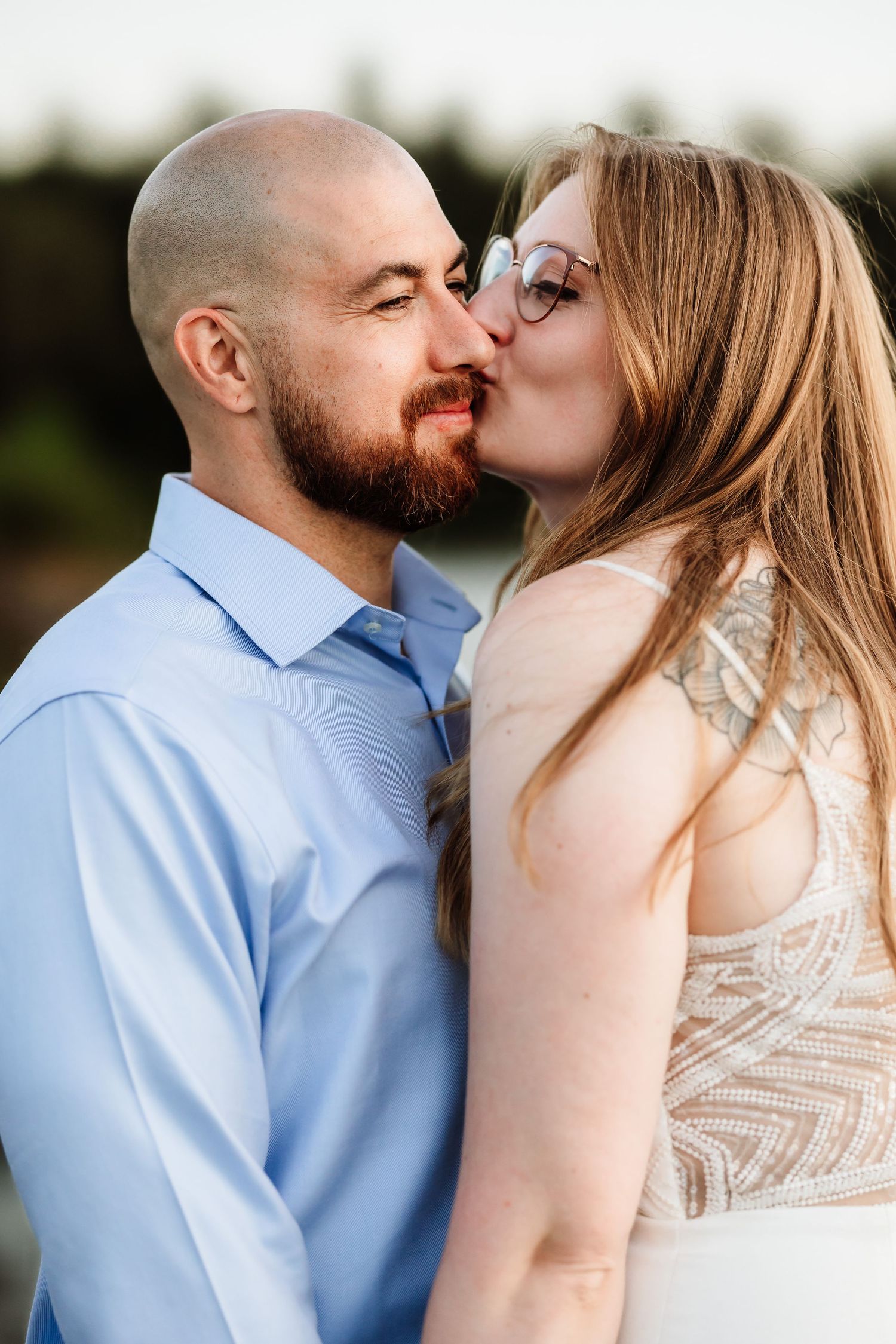 Bald bearded man in blue shirt shares a romantic kiss with long-haired woman wearing glasses outdoors.