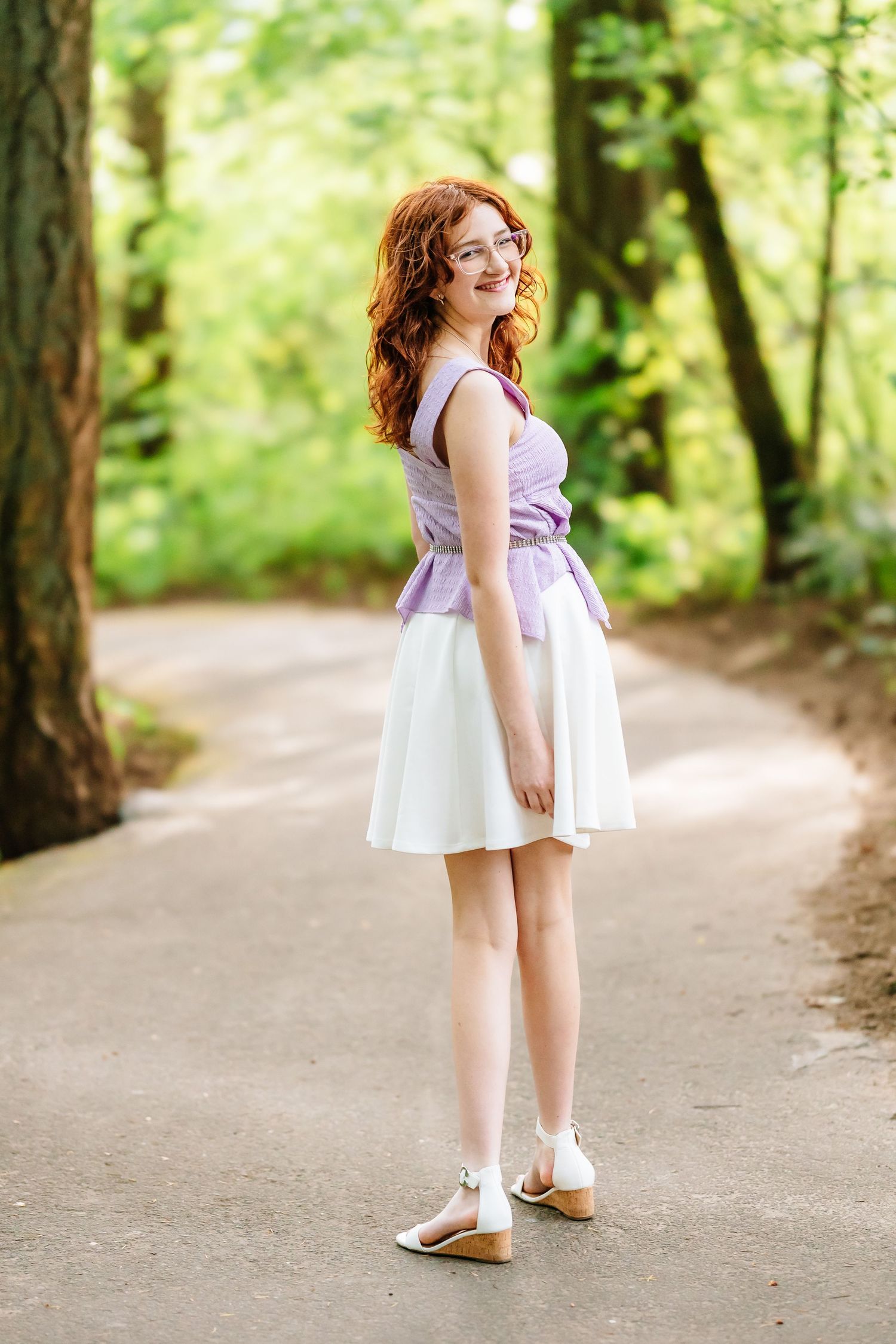 Young redhead woman in purple top and white skirt smiling on a tree-lined path in a lush green forest.