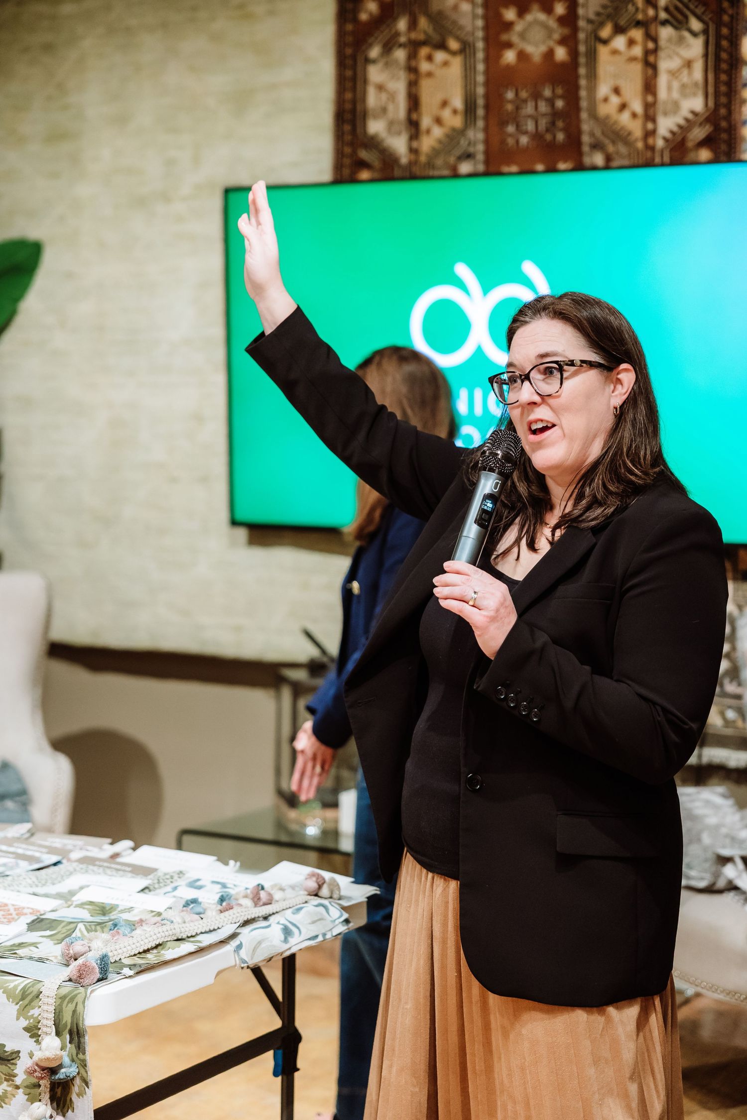Woman in black blazer speaking into microphone with hand raised at a design event.
