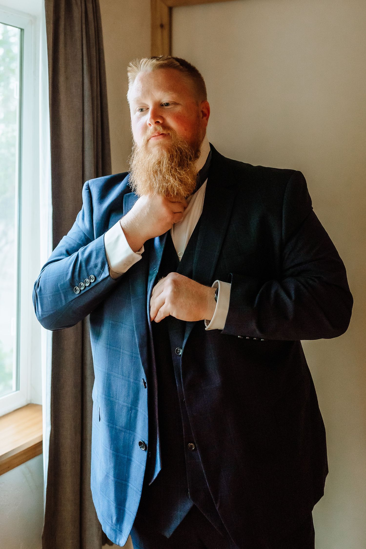 Bearded groom in a blue suit adjusting his tie while getting ready by a window.
