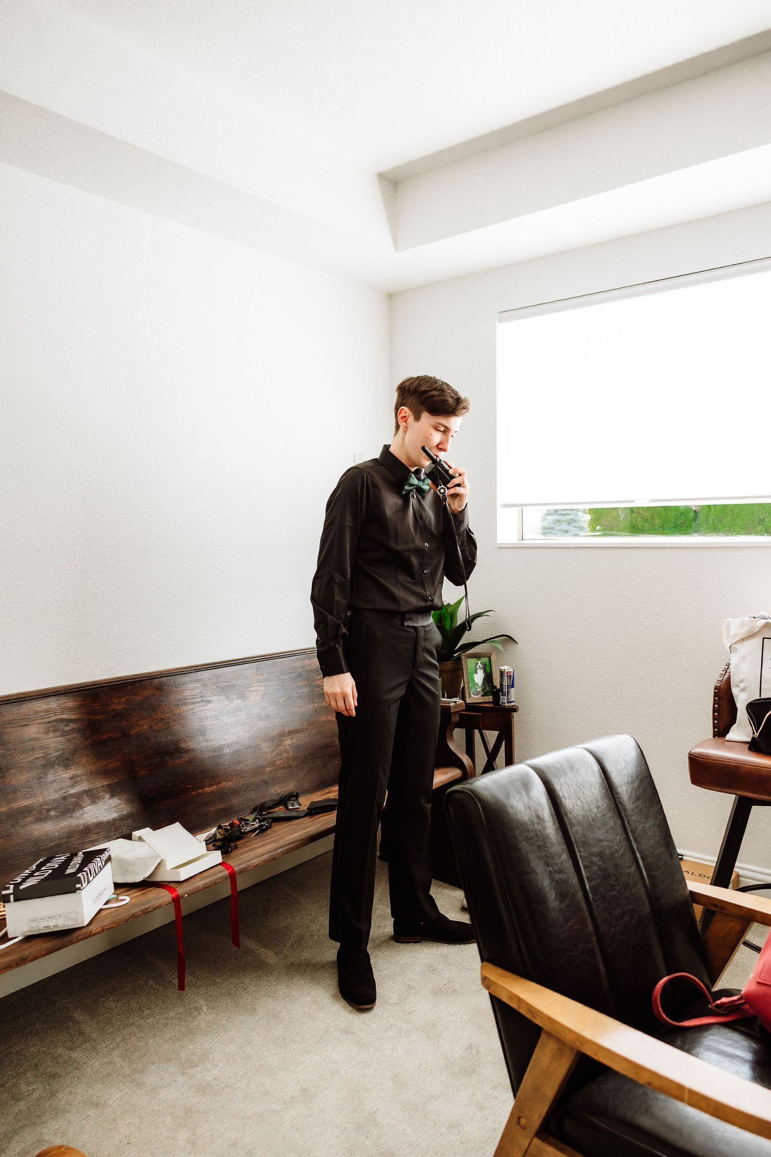 Groom in black shirt and trousers talks on phone while standing by a bright window.