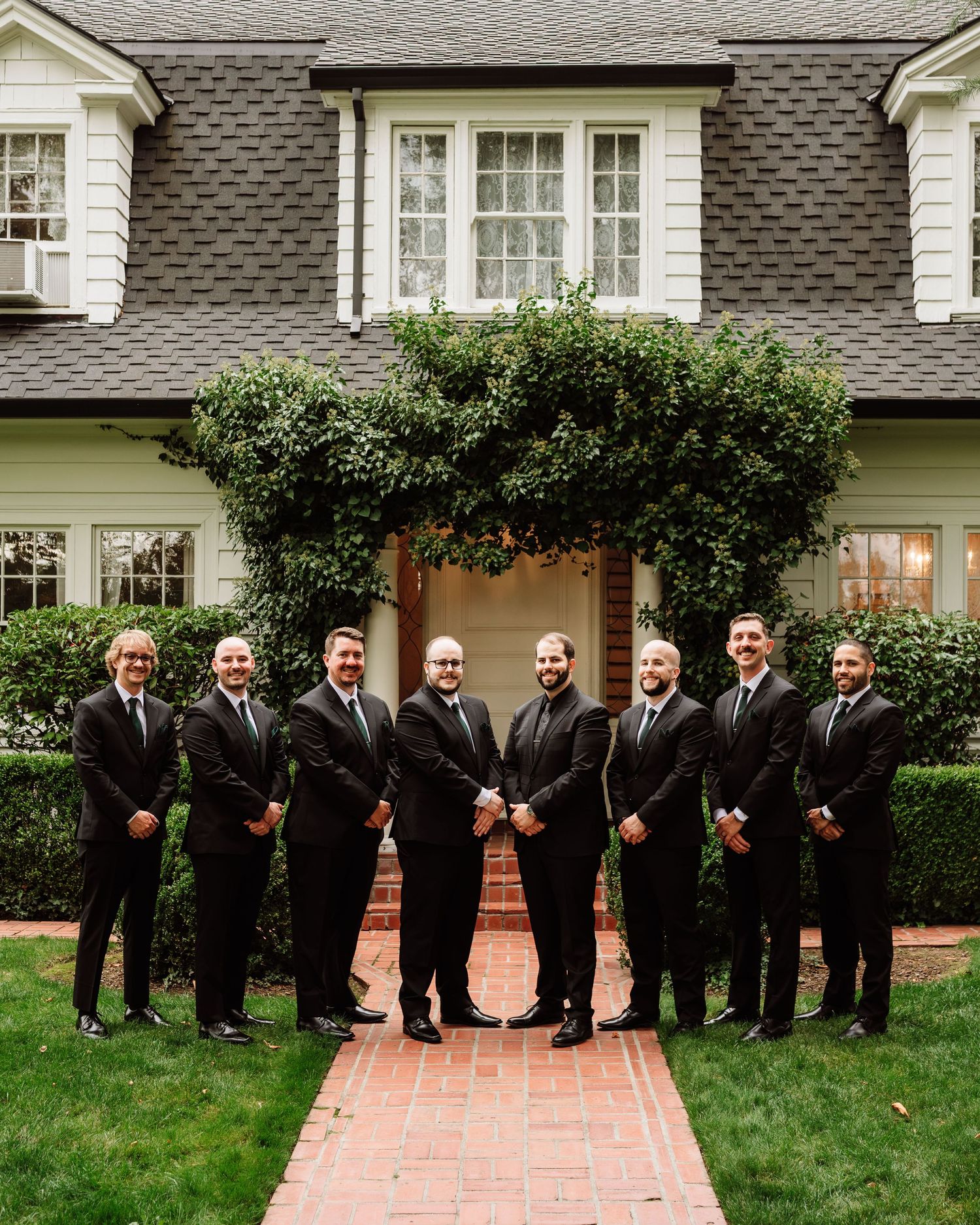 Eight groomsmen in black suits pose symmetrically on a brick path in front of an elegant house.