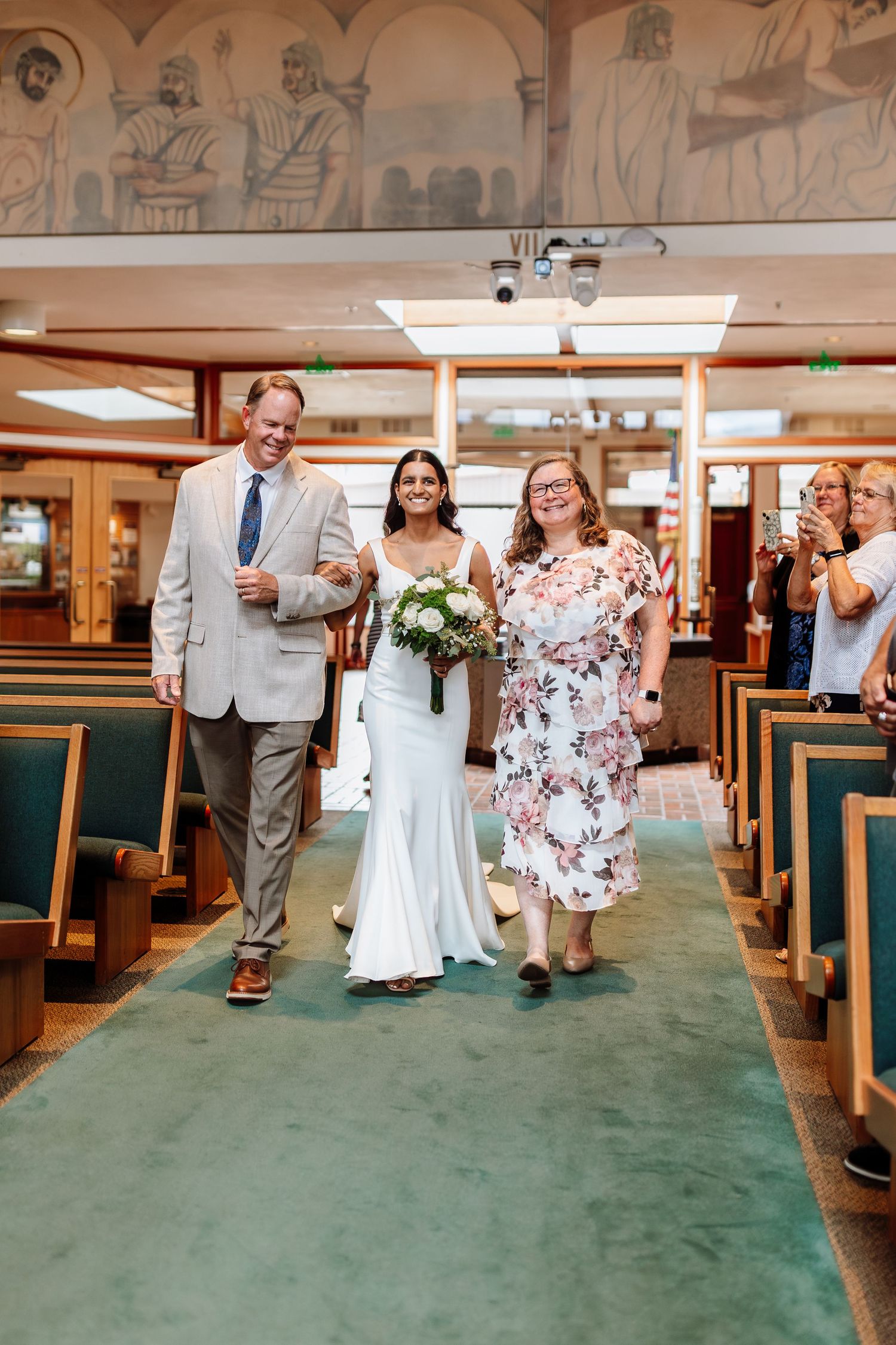 Bride in white gown walks down church aisle escorted by two family members holding bouquet.