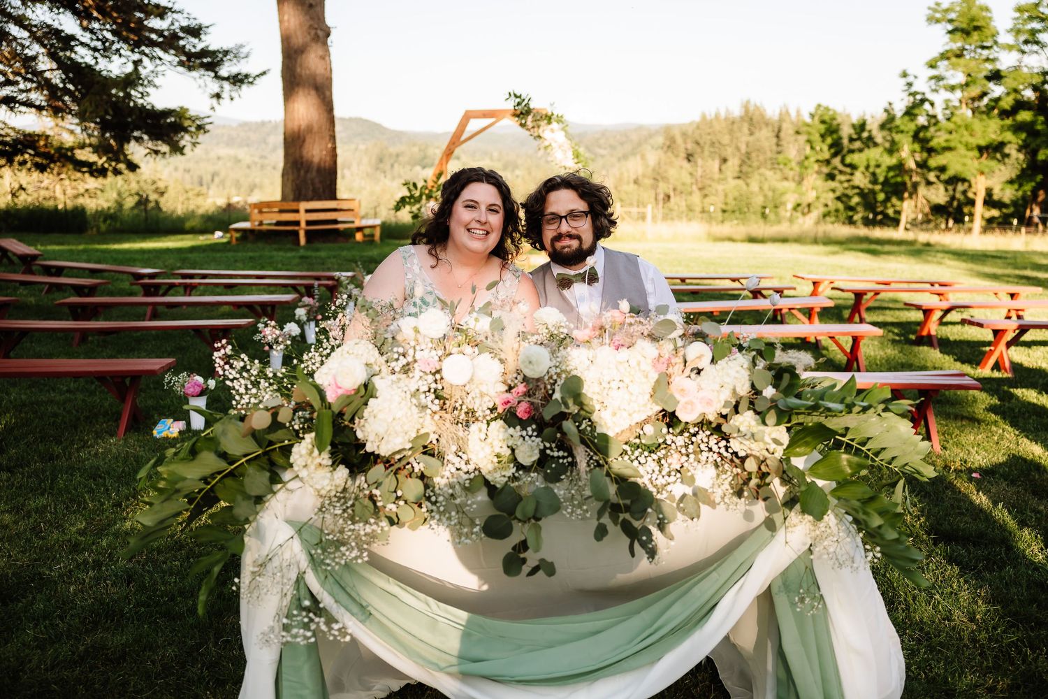 Bride and groom smile at sweetheart table adorned with lush white floral arrangements outdoors.