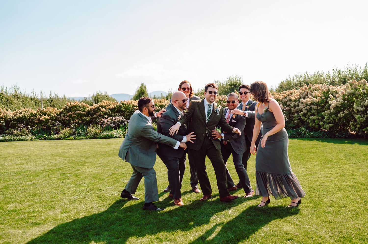 Groom and groomsmen pose playfully wearing sunglasses on a bright sunny lawn at a wedding.