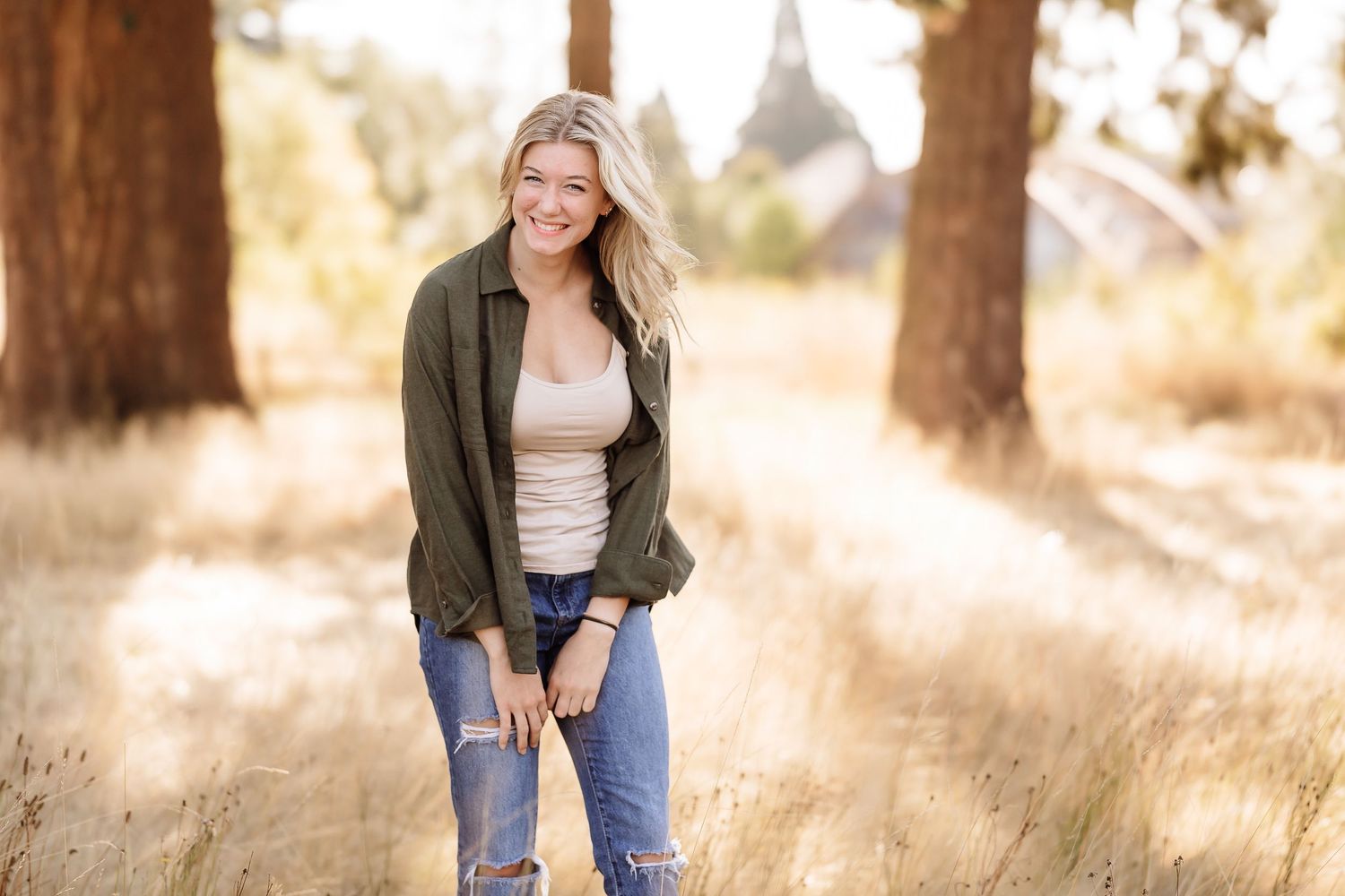 Young woman in olive jacket and ripped jeans smiling in sunlit forest clearing.