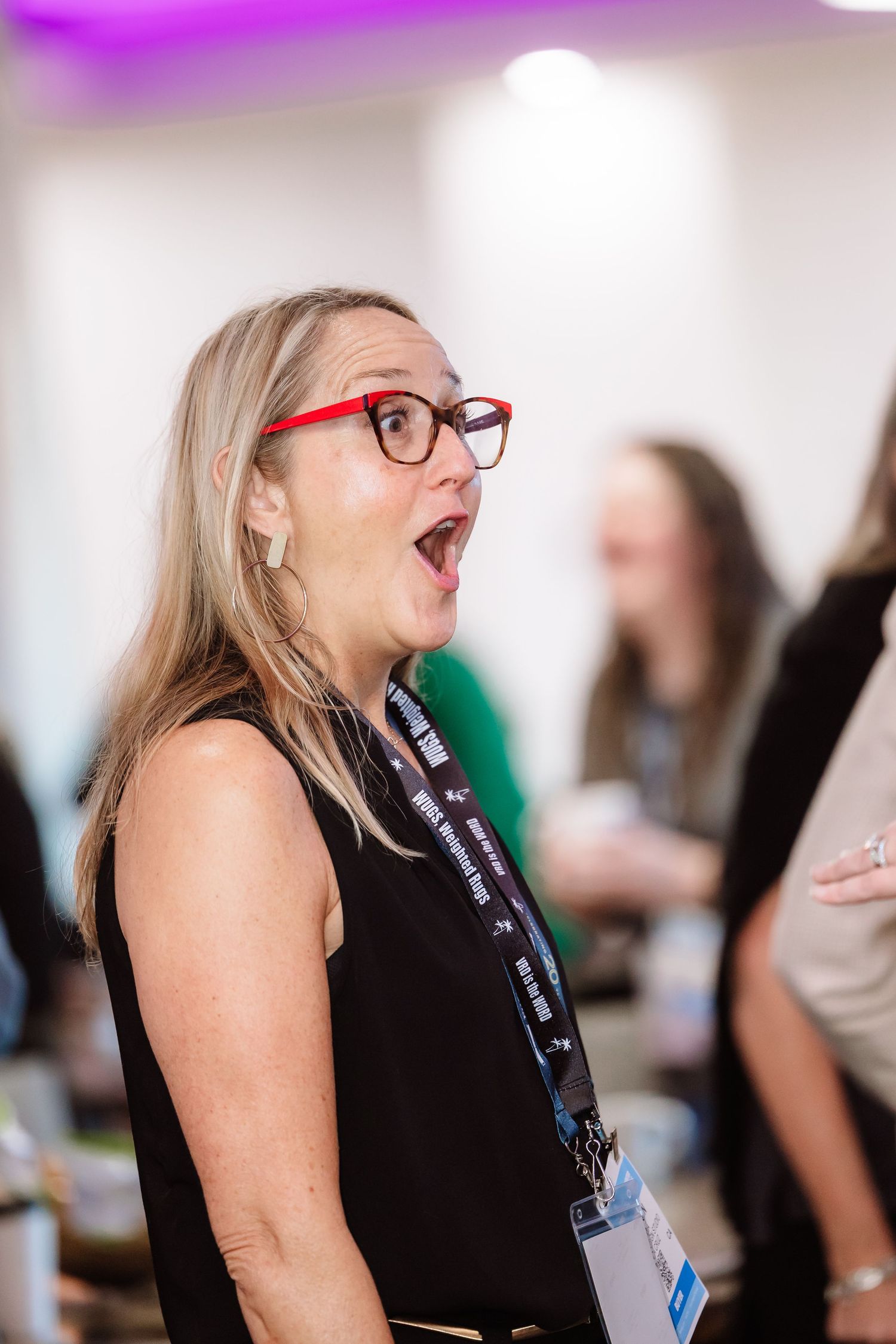 Blonde woman with red glasses reacting with surprised expression at a networking event.