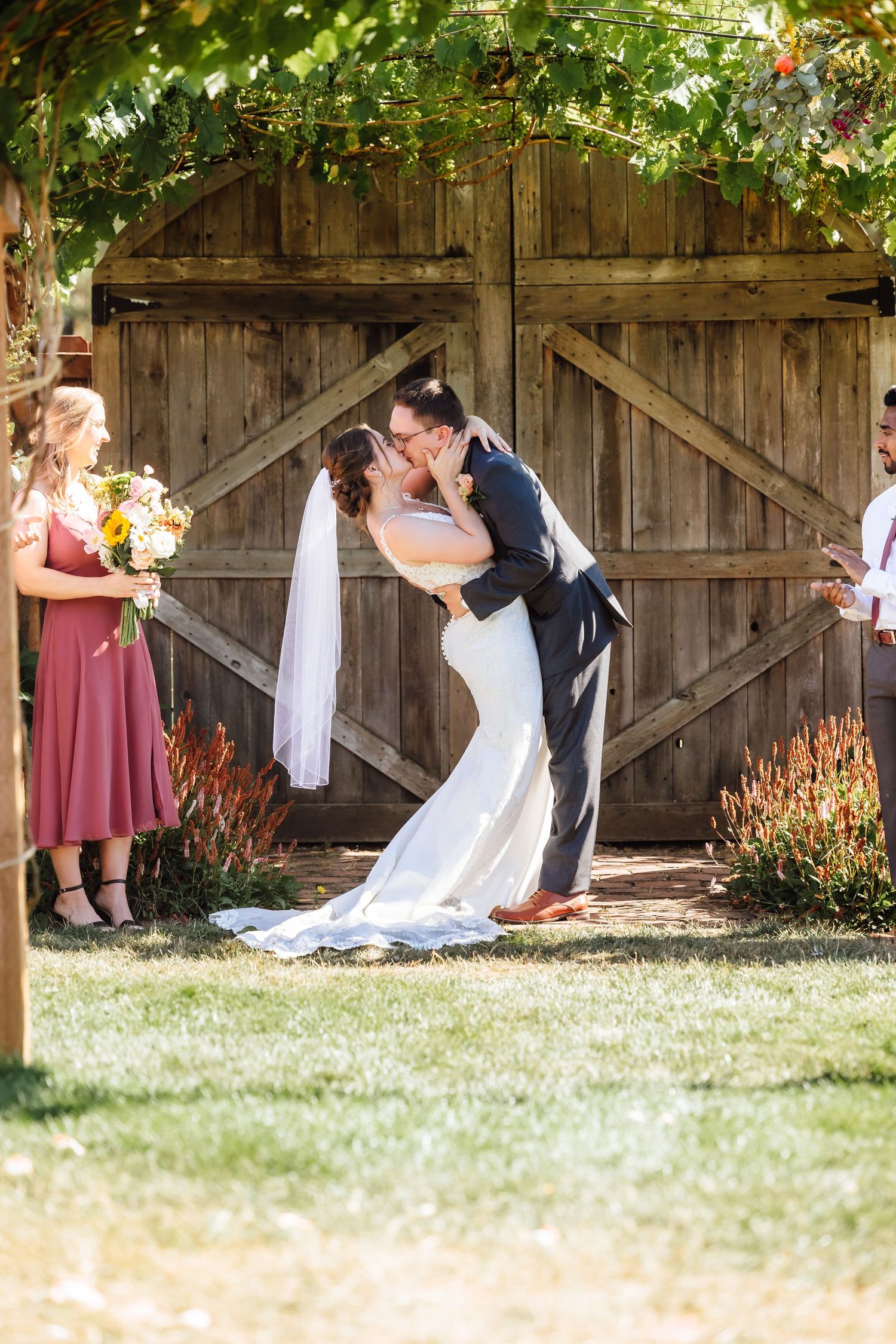 Groom dips bride for romantic first kiss at rustic barn outdoor wedding ceremony with wedding party.