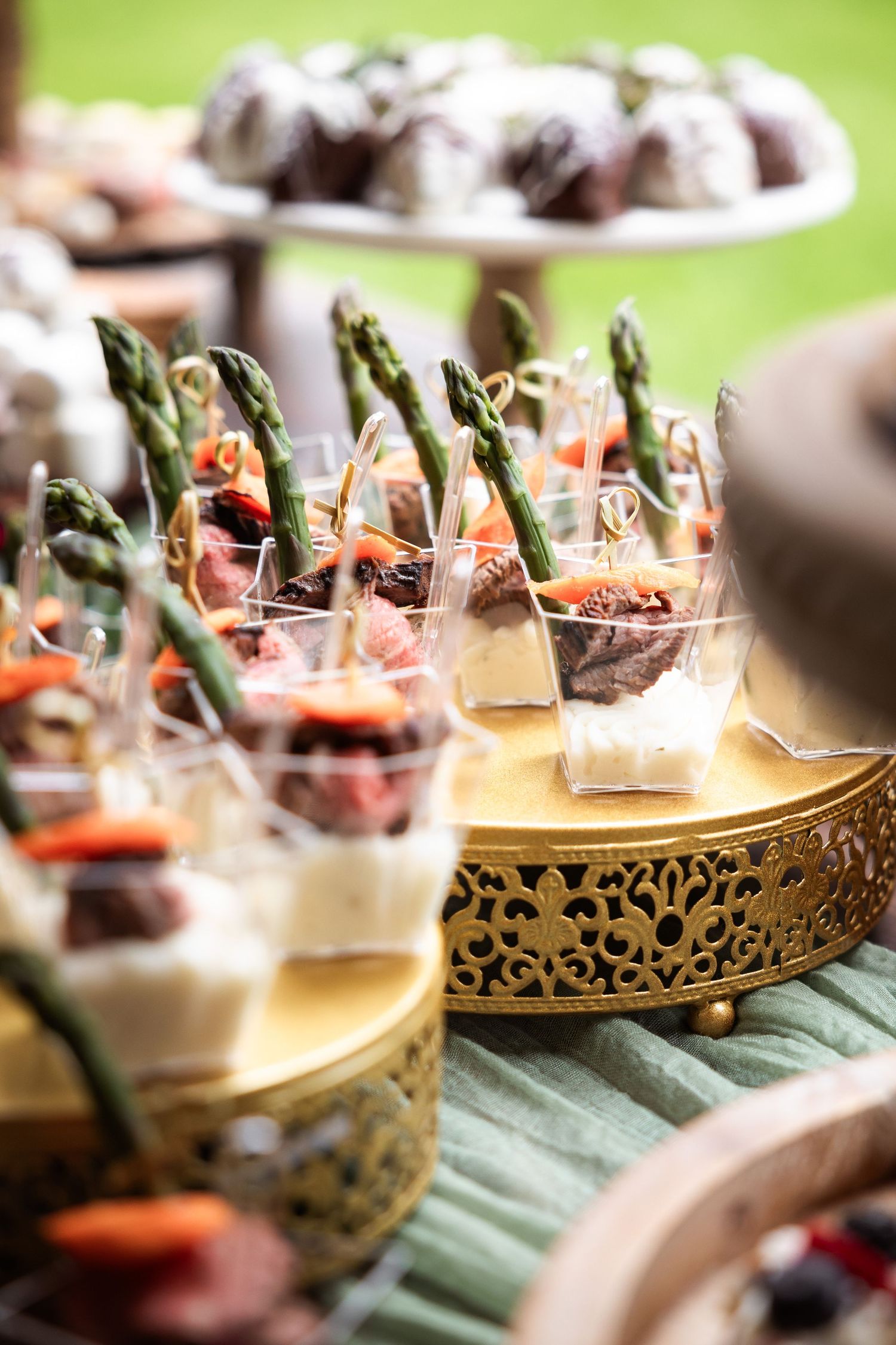 Elegant wedding appetizer display featuring asparagus, charcuterie cups, and chocolate truffles on ornate gold trays.