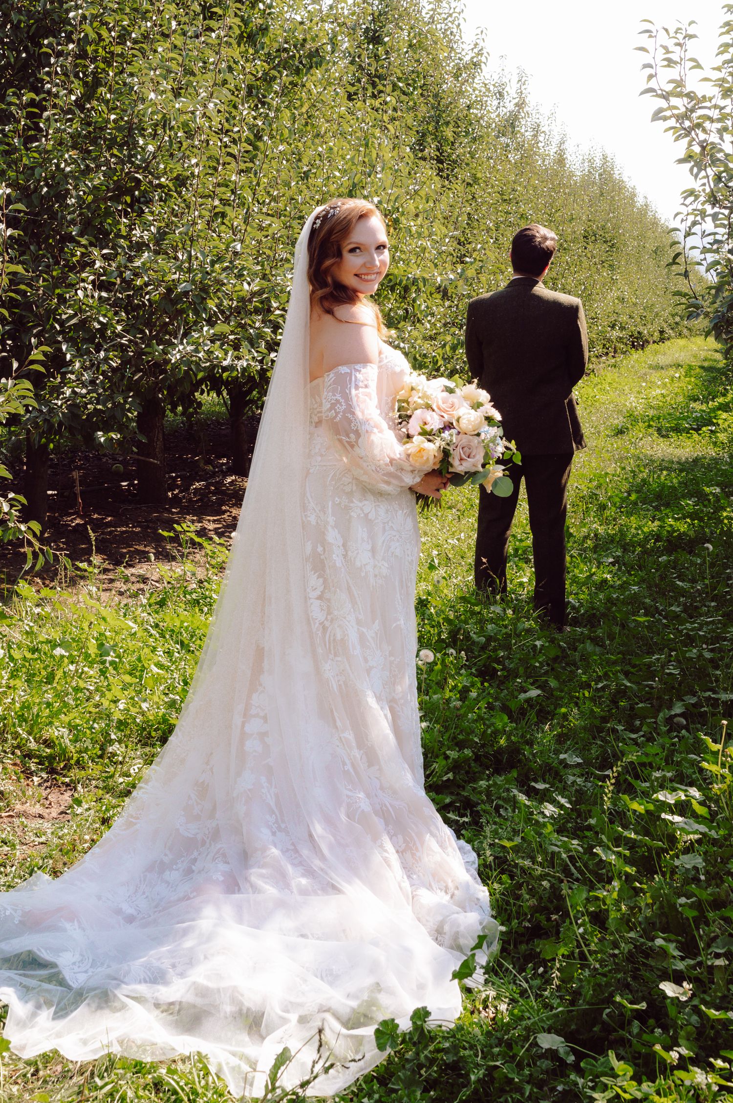 Bride in lace gown with veil and bouquet poses in sunlit orchard as groom walks ahead.
