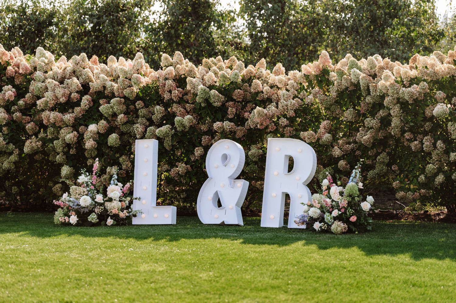 Large white marquee letters L and R on lawn with blooming hydrangea hedge and floral arrangements.