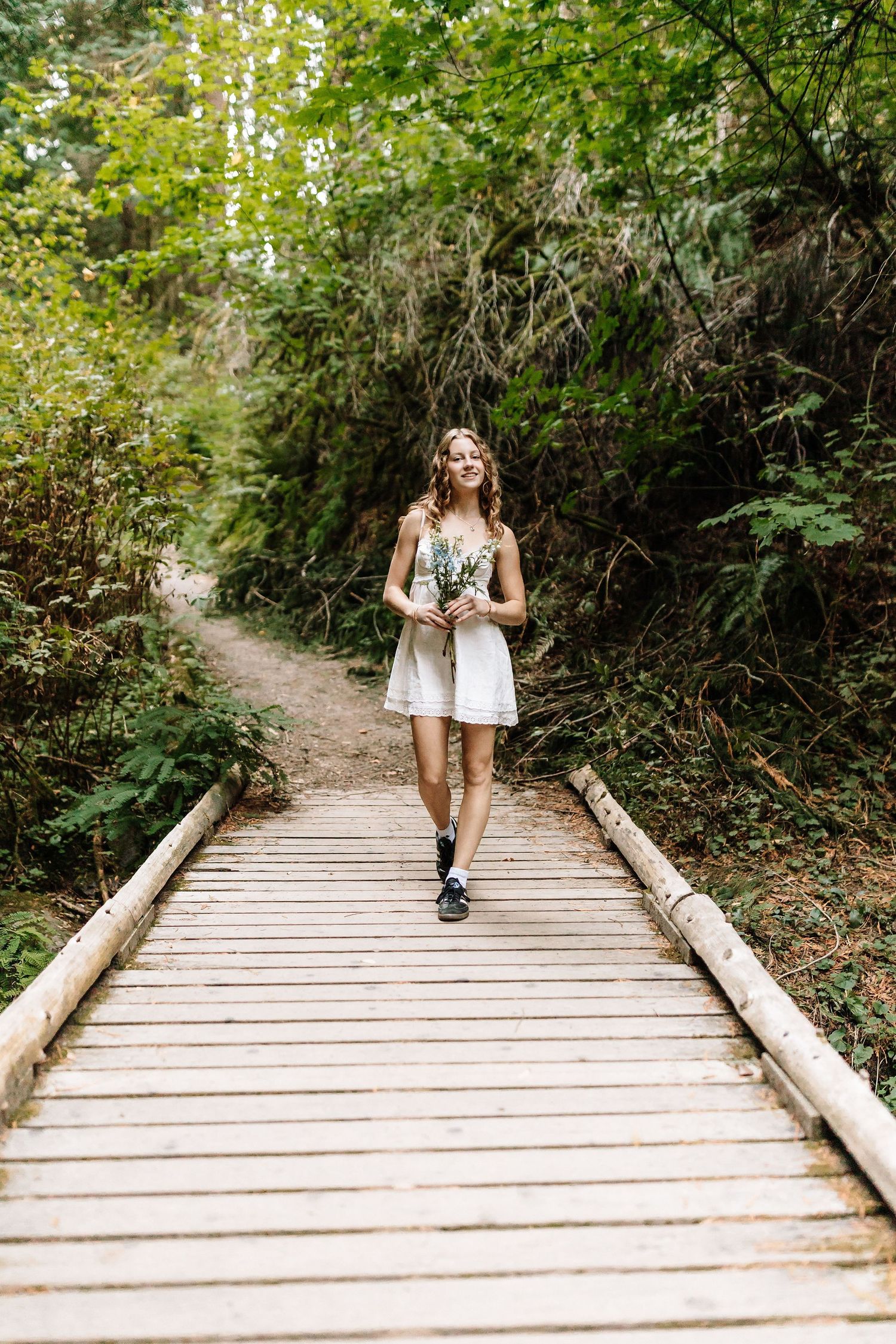 Young woman in white dress holding wildflowers walks along a wooden boardwalk path through a lush green forest.