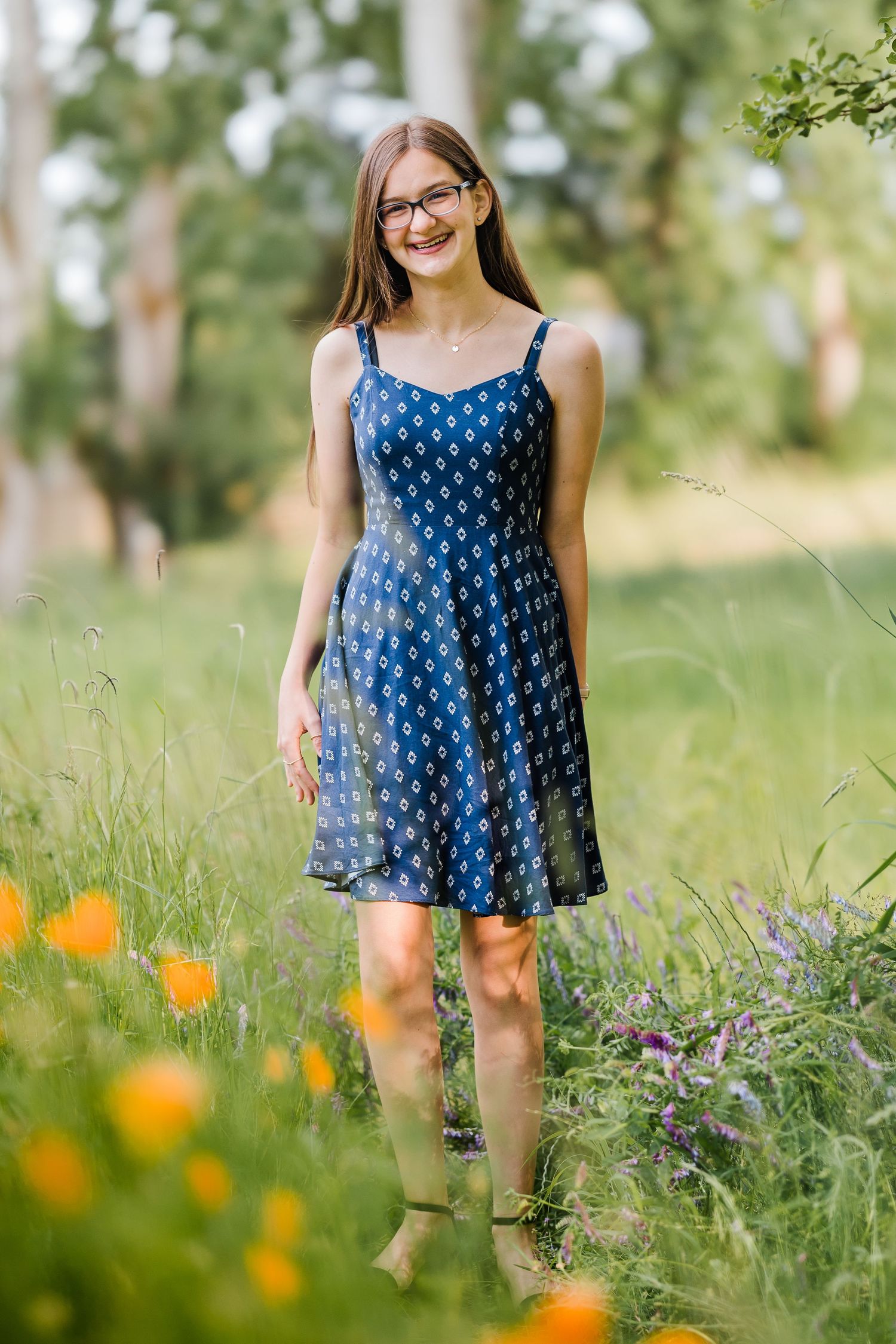 Smiling teenage girl in blue patterned sundress standing in a wildflower meadow with orange and purple blooms.
