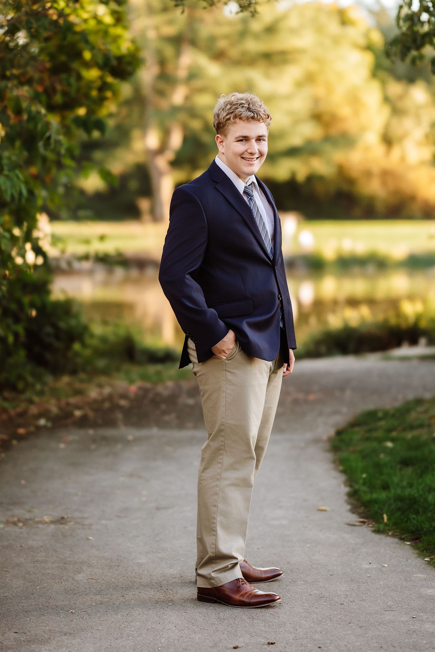 Smiling teenage boy in navy blazer, tie, and khakis poses on a park path near a pond with autumn trees.