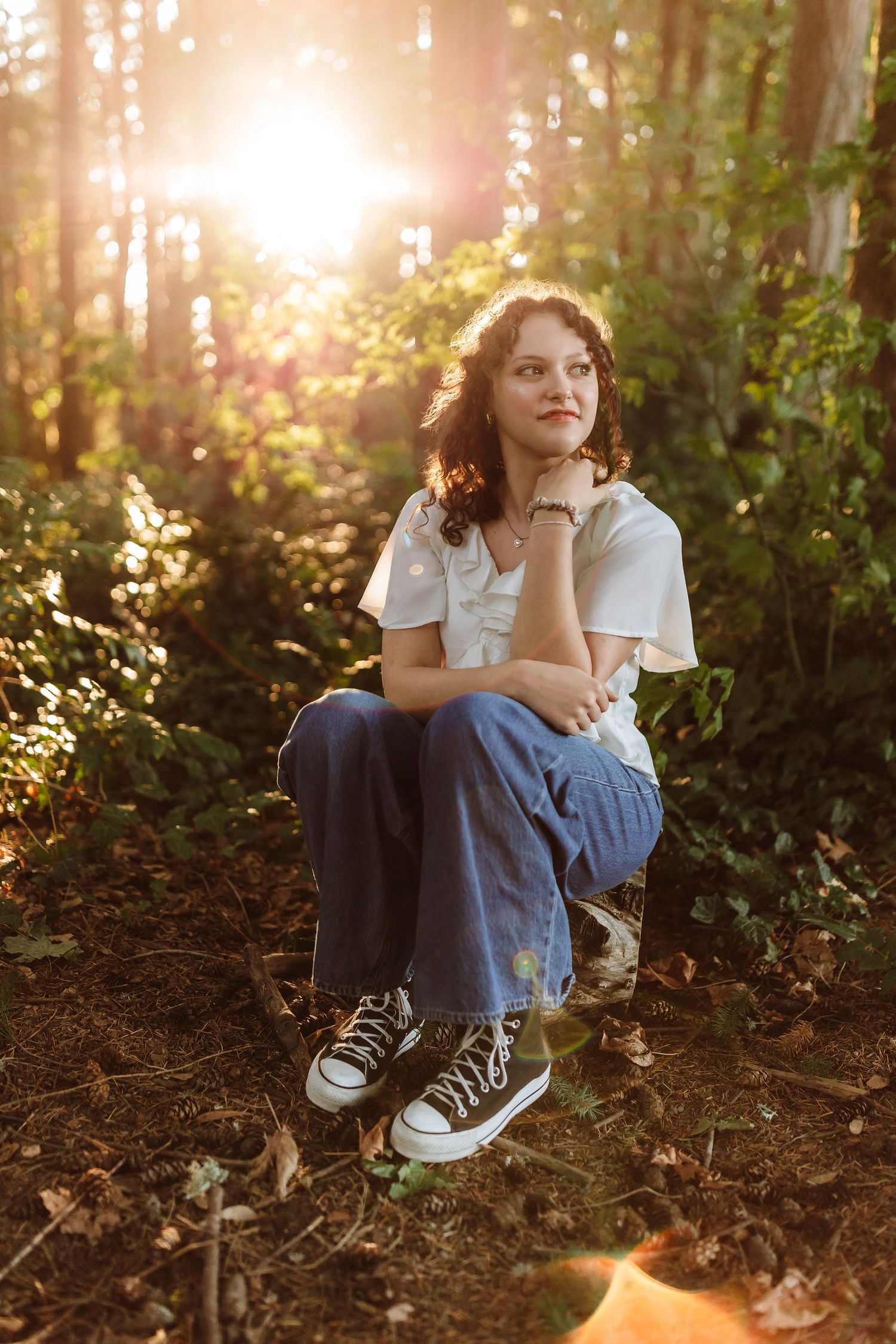 Young woman in white top and jeans sits in sunlit forest, golden light streaming through trees behind her.