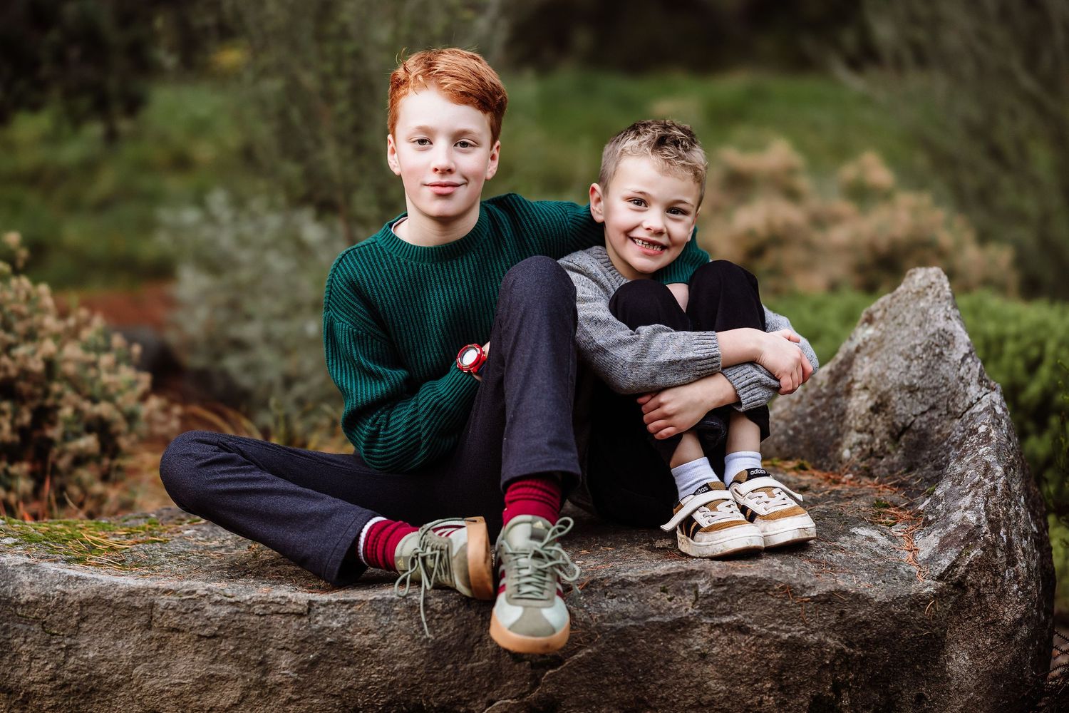 Two boys sitting on a rock outdoors, smiling, one with red hair in green sweater, younger in gray outfit.