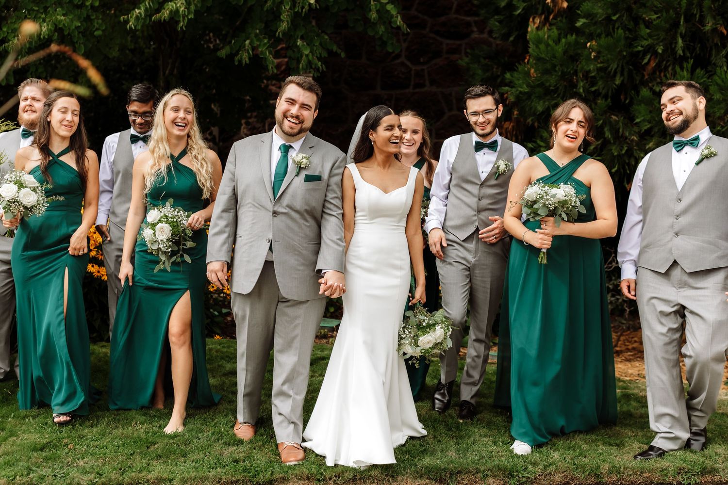 A bride and groom walk hand in hand with their wedding party in green and gray attire outdoors.