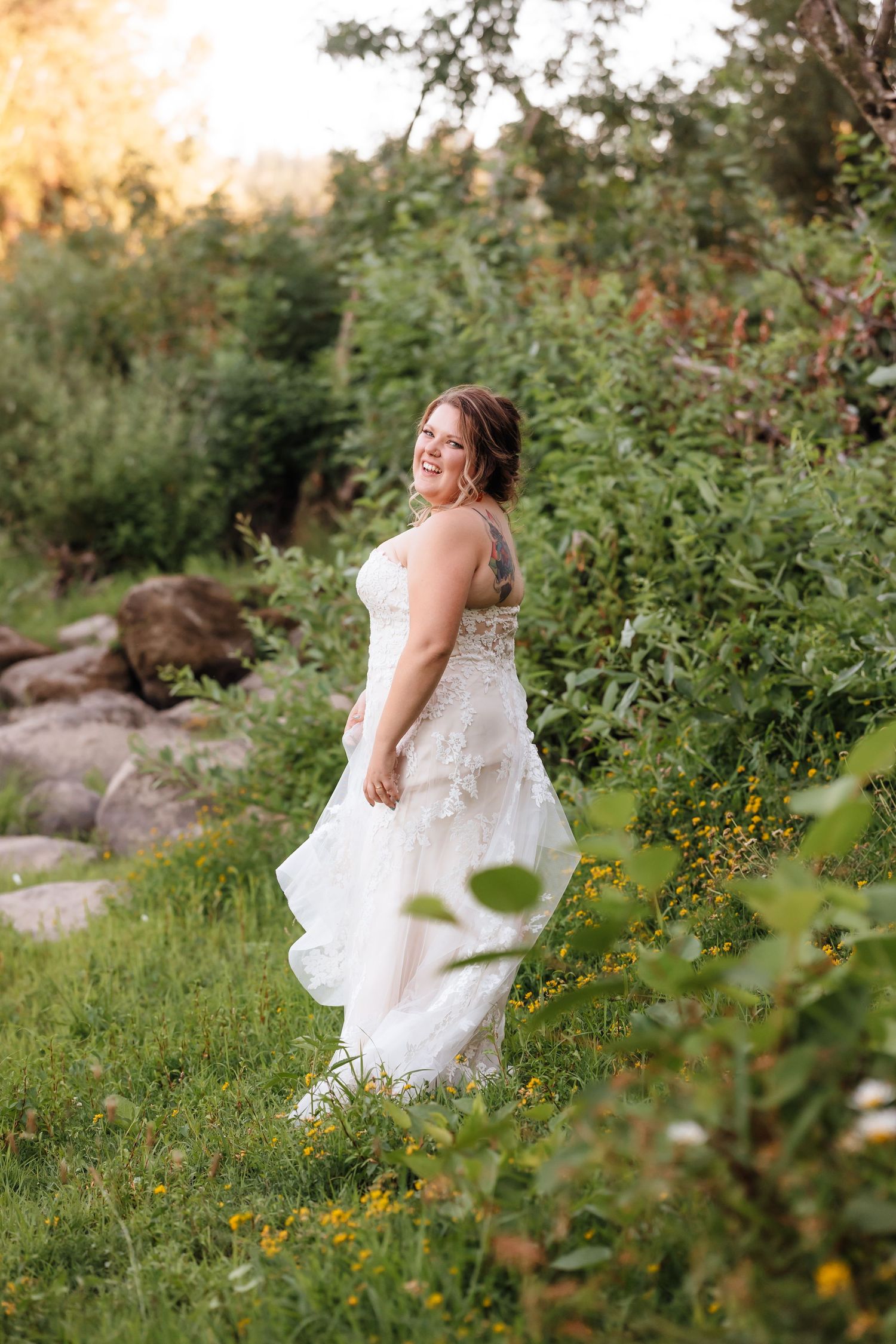 Radiant bride in a lace wedding gown twirling joyfully among wildflowers and greenery in golden evening light.