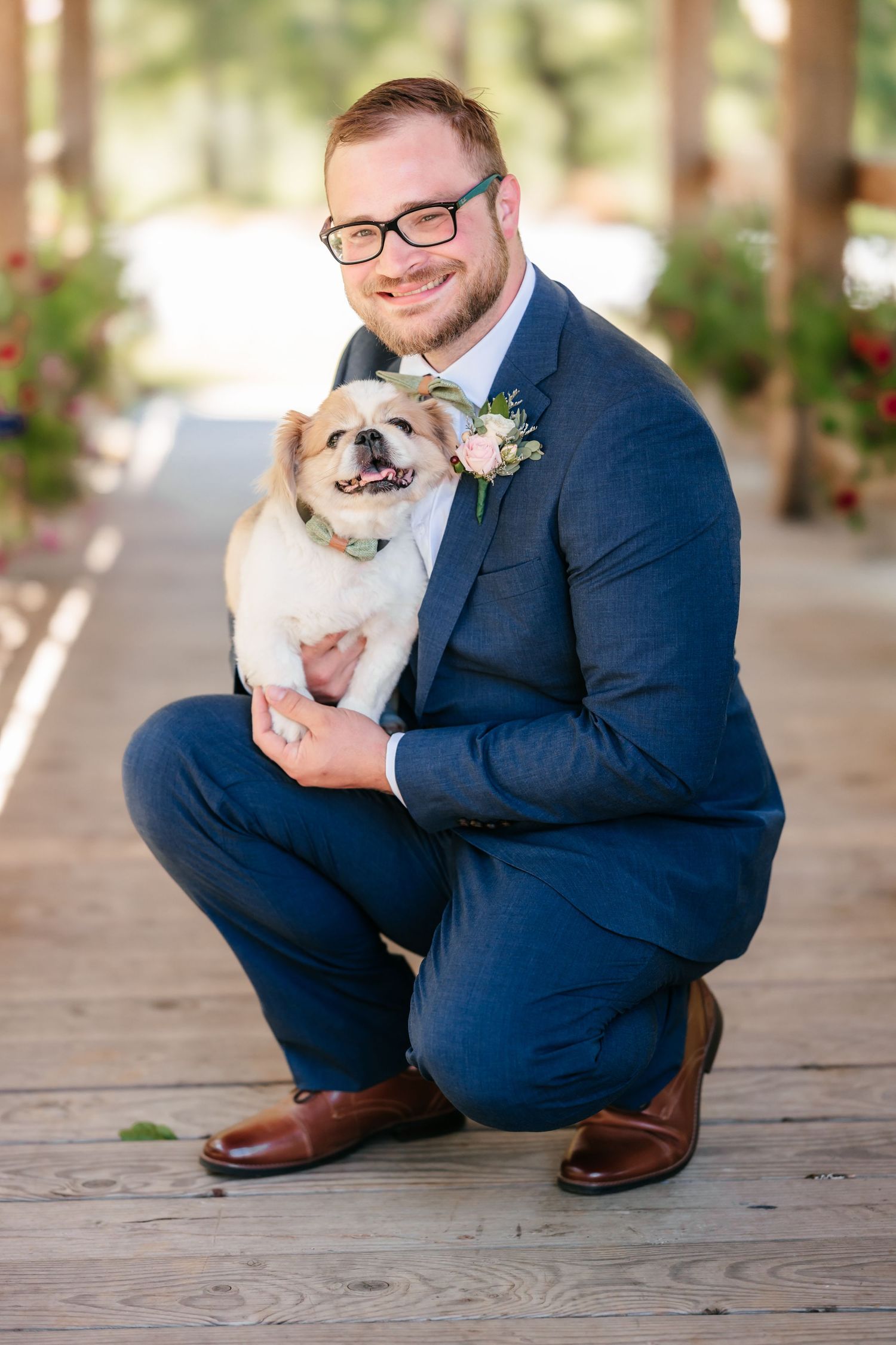 Groom in navy suit crouching down while holding a small fluffy dog wearing a boutonniere on a wooden deck.