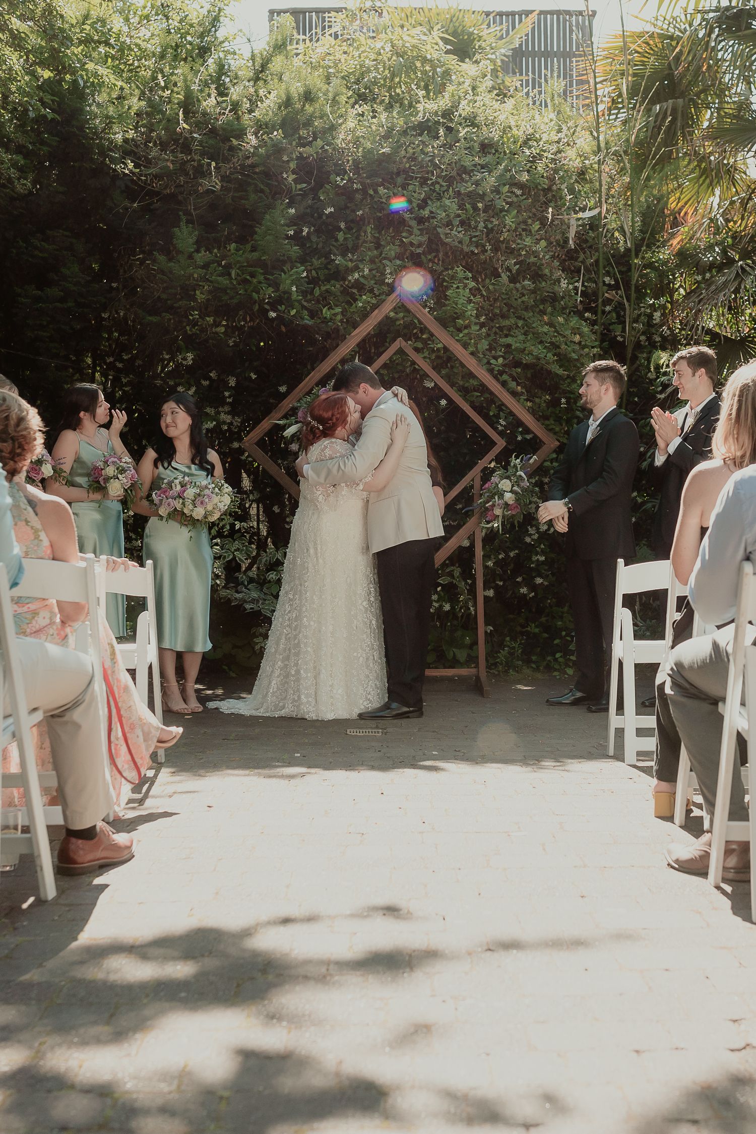 Newlyweds share a kiss at the altar under a geometric wooden arch surrounded by wedding party.