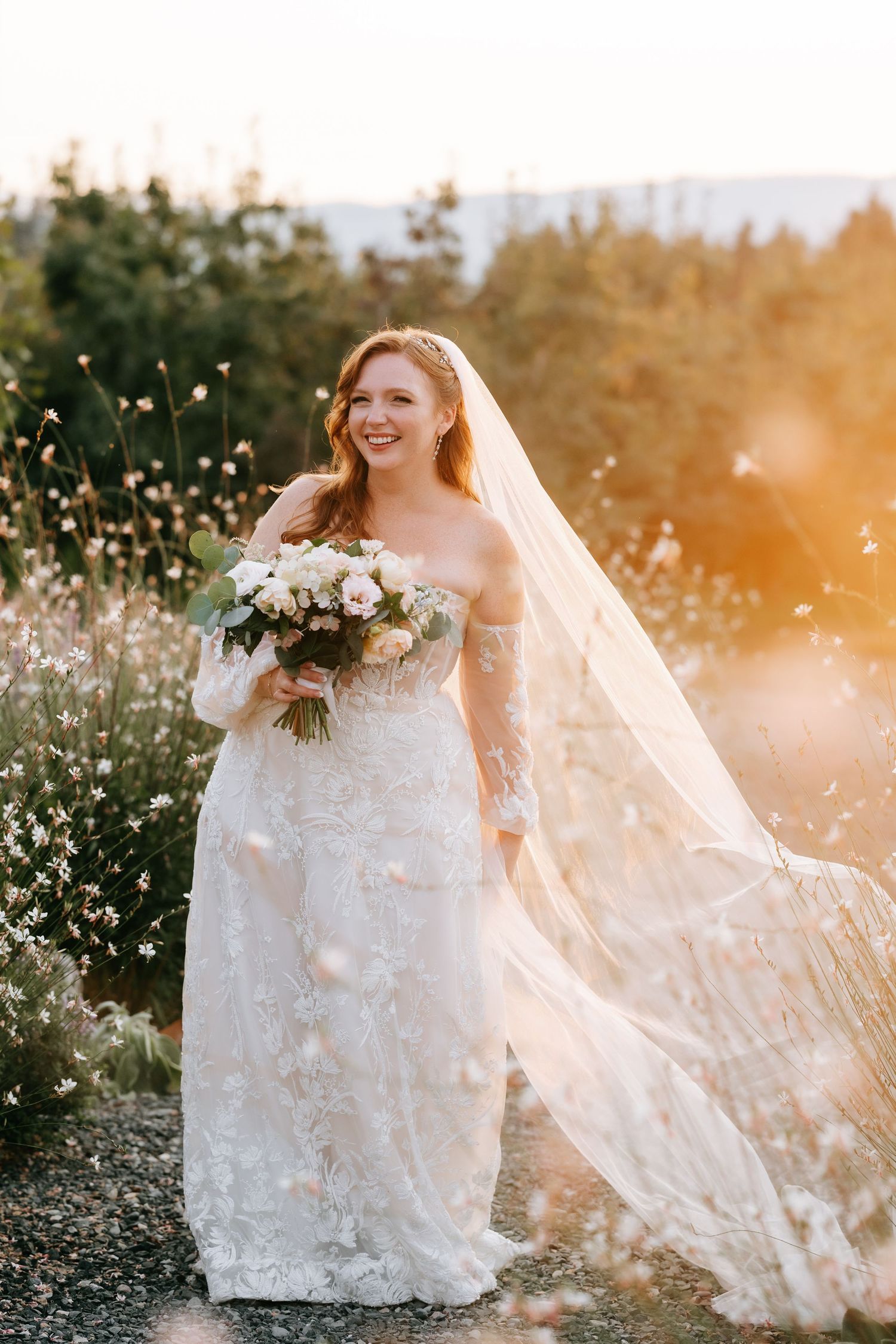 Radiant bride in lace gown and flowing veil holds bouquet in golden sunset light among wildflowers.