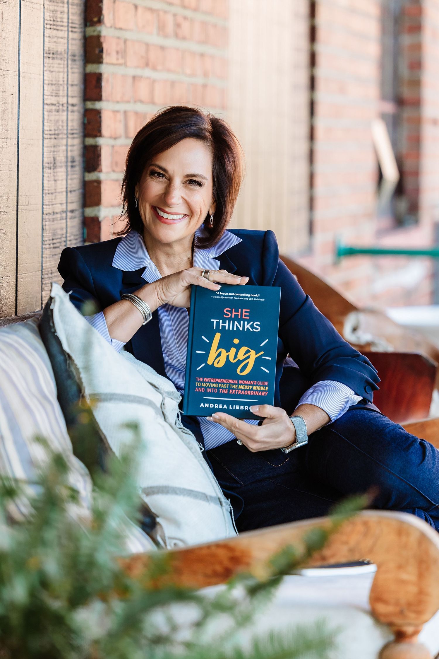 Smiling woman in blue blazer holding She Thinks Big book while seated on cozy porch bench.