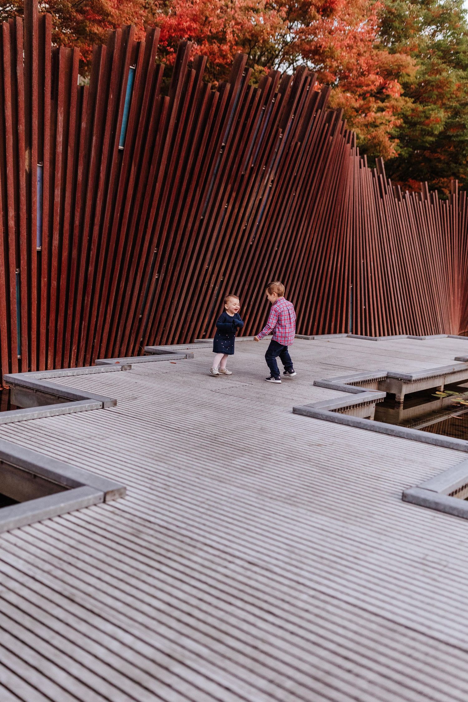 Two young children playing on a modern concrete deck beside tall russet metal poles with autumn foliage in the background.