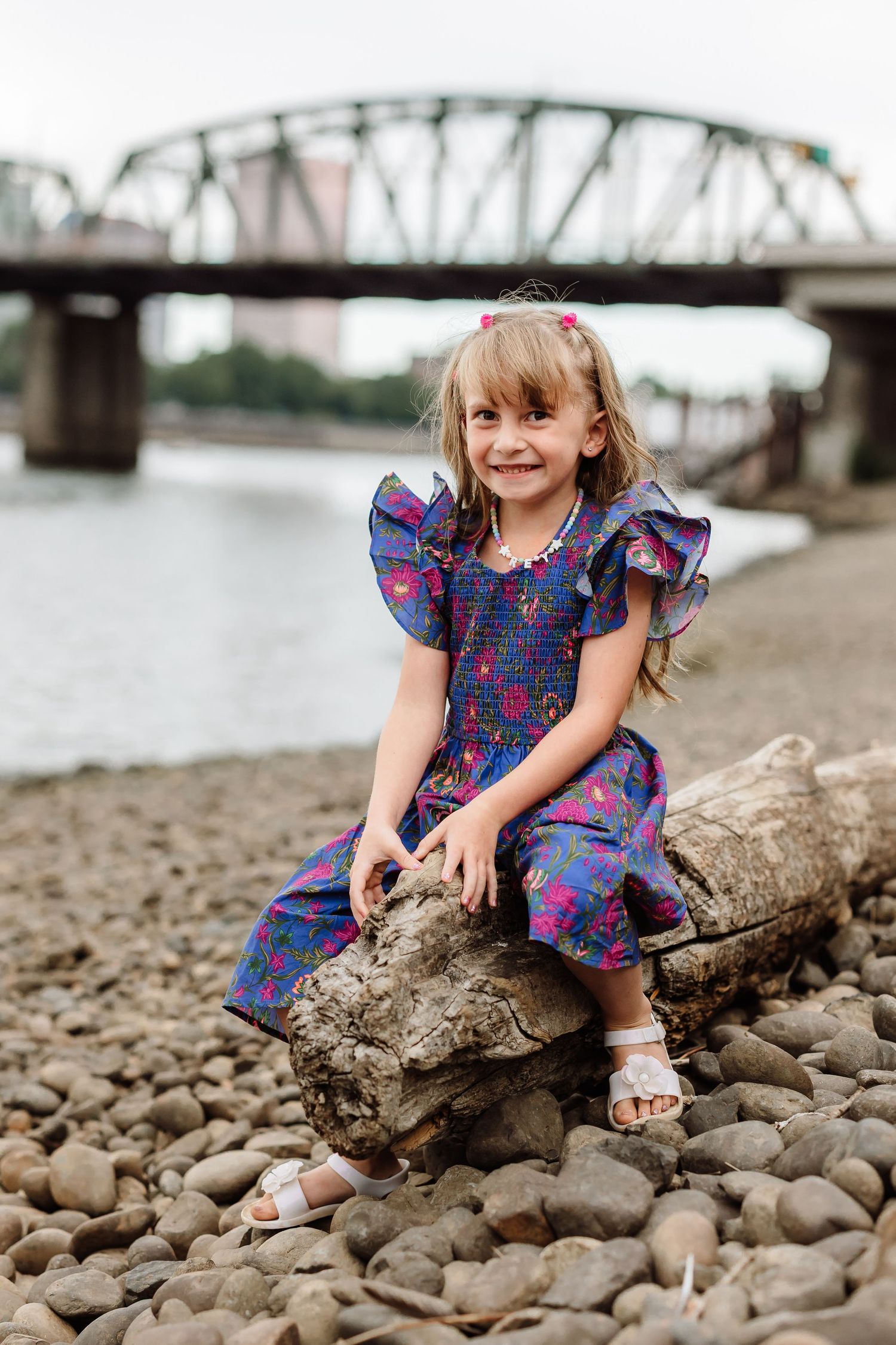 Young girl in blue floral dress sitting on driftwood by rocky riverbank with bridge in background.