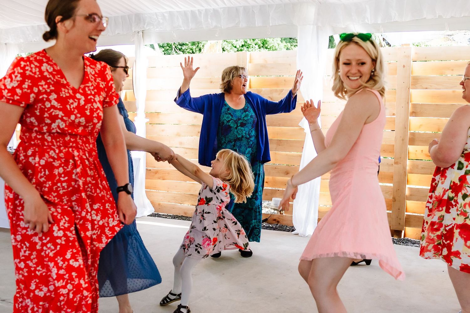 Women and a little girl dancing joyfully at a wedding reception under a white tent with wooden backdrop.