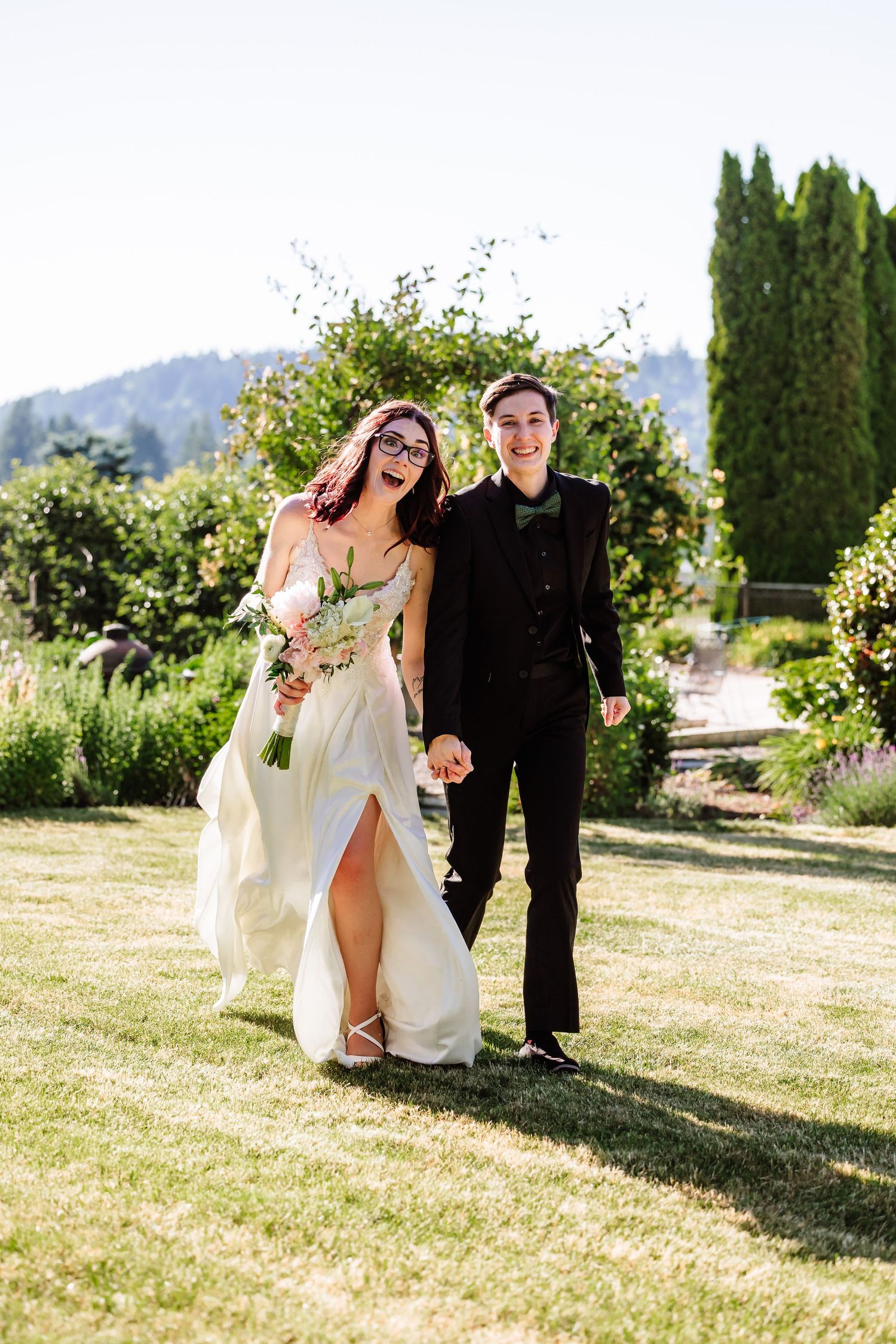 Laughing bride in white gown and partner in black suit walk through a lush garden.