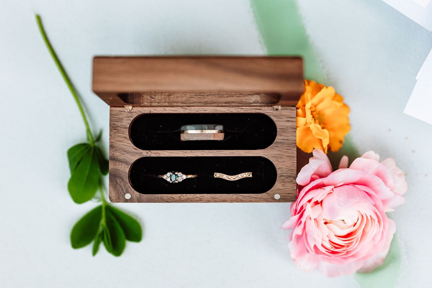 Wedding rings displayed in a wooden velvet-lined ring box with pink and orange roses nearby.