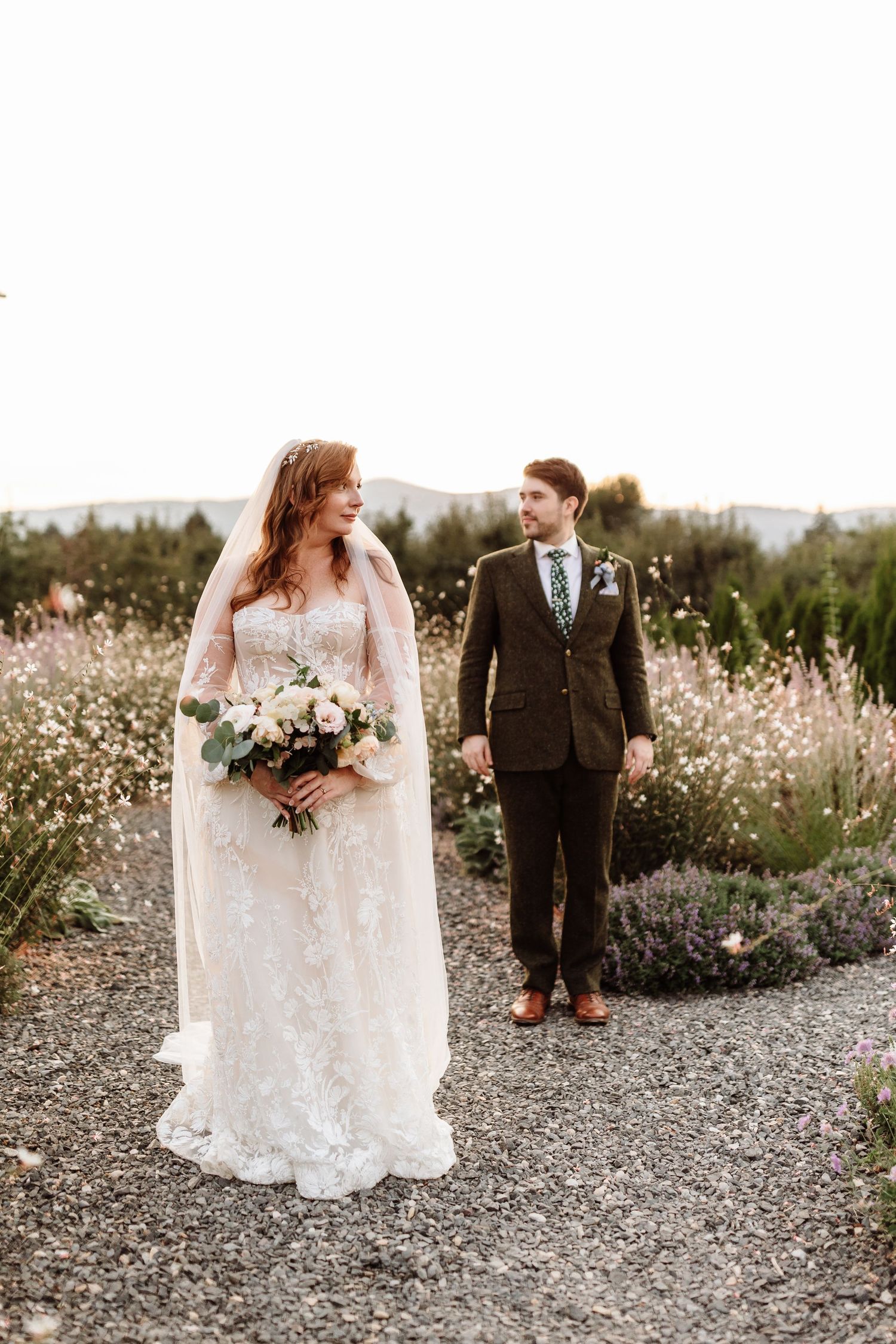 Bride and groom hold hands on garden path surrounded by wildflowers in soft natural light.