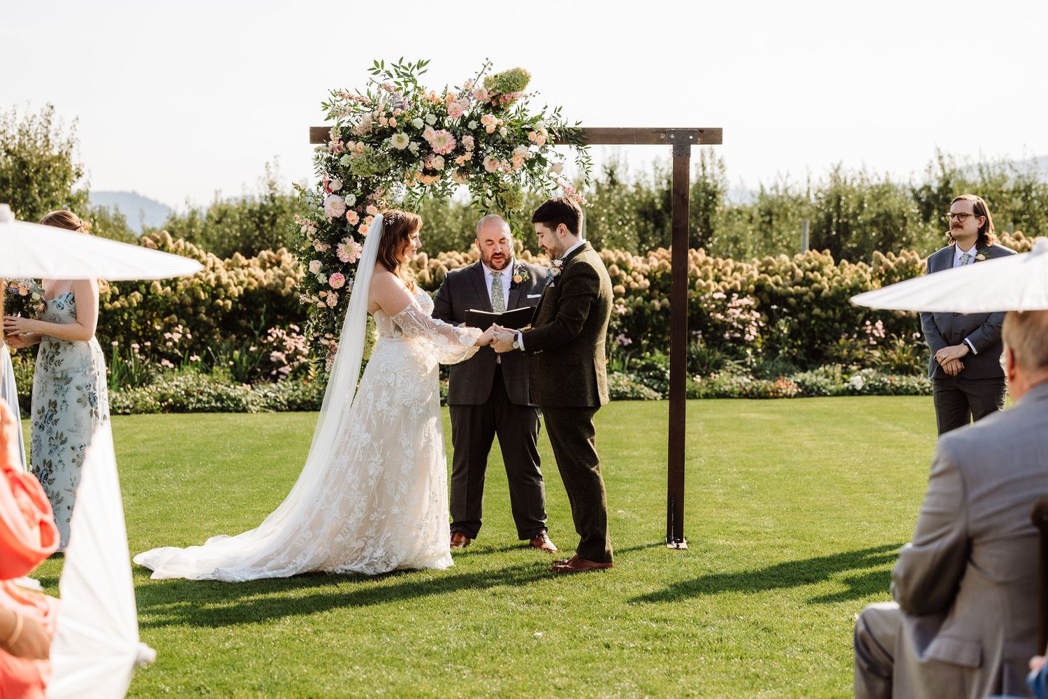 Bride and groom exchange vows at an outdoor wedding ceremony with a floral arch backdrop.