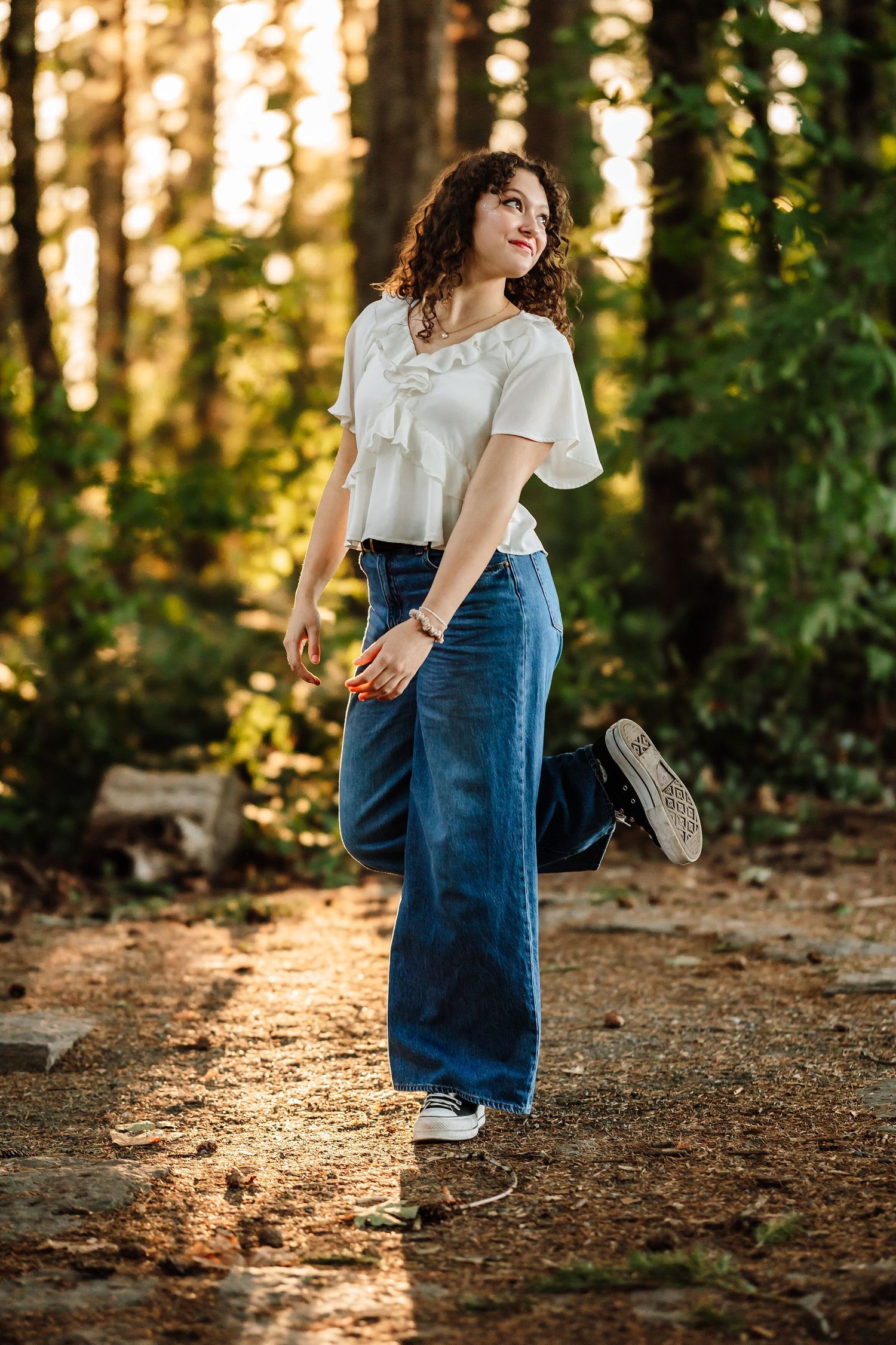 Woman in white top and wide-leg jeans poses playfully on a sunlit forest path surrounded by tall trees.