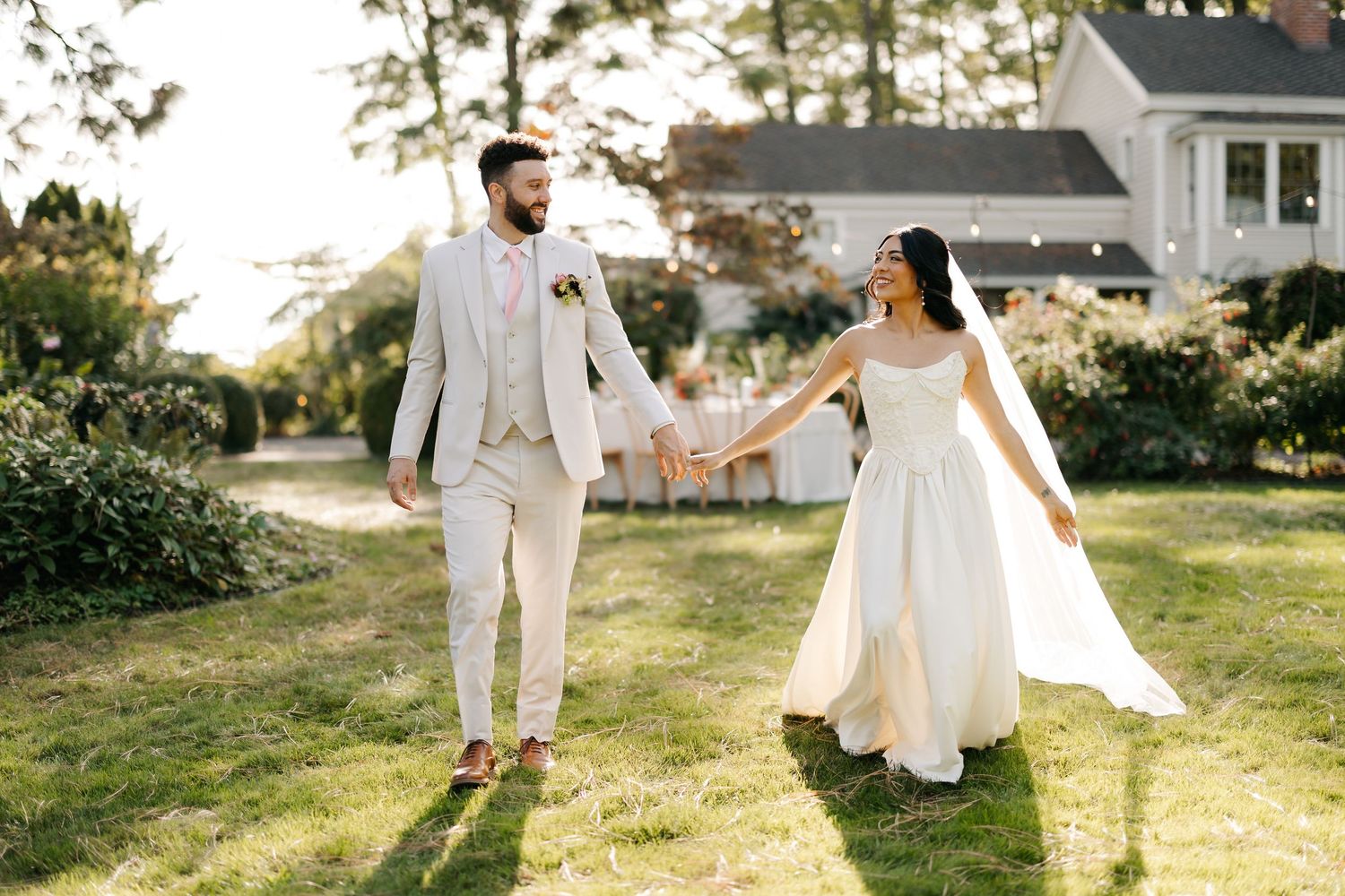 Bride and groom holding hands walking through a sunlit garden near a charming white house.