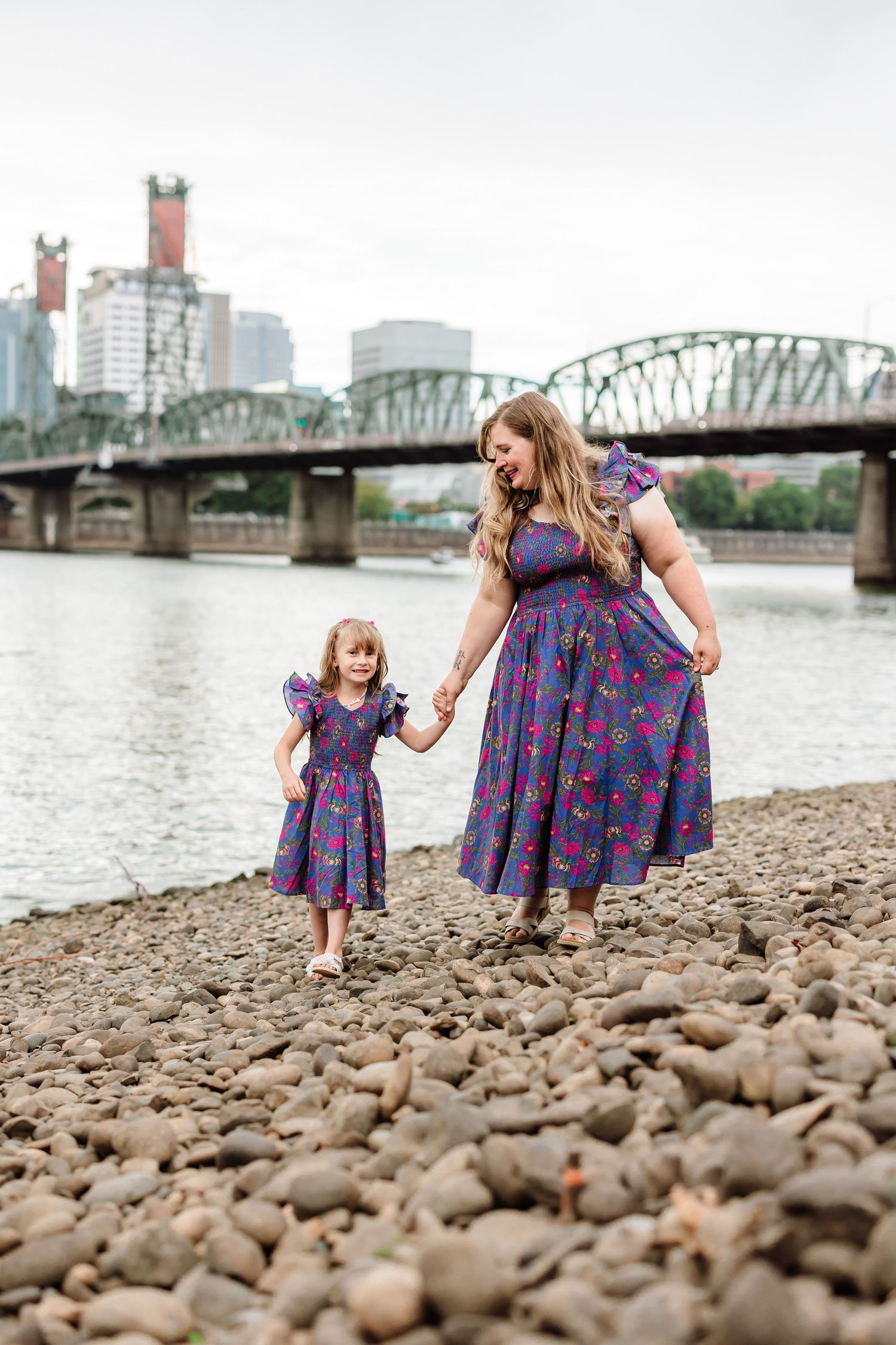 Mother and daughter in matching purple floral dresses hold hands on rocky riverbank with bridge and city skyline behind them.