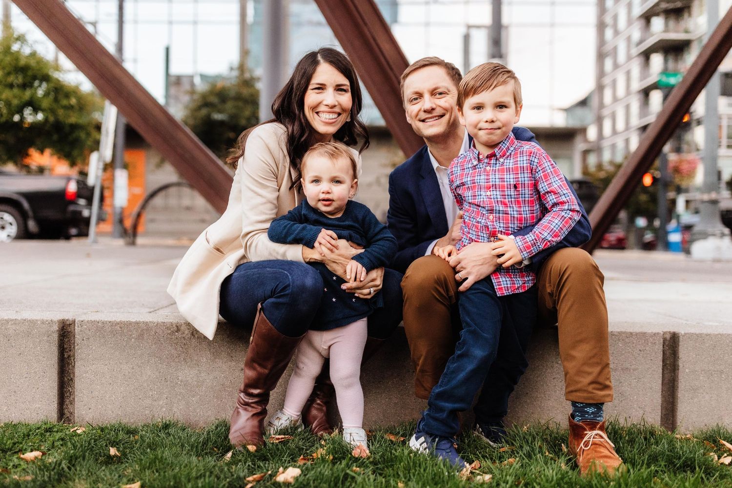 Happy family of four posing outdoors in urban setting, parents smiling with toddler girl and young boy on stone bench.