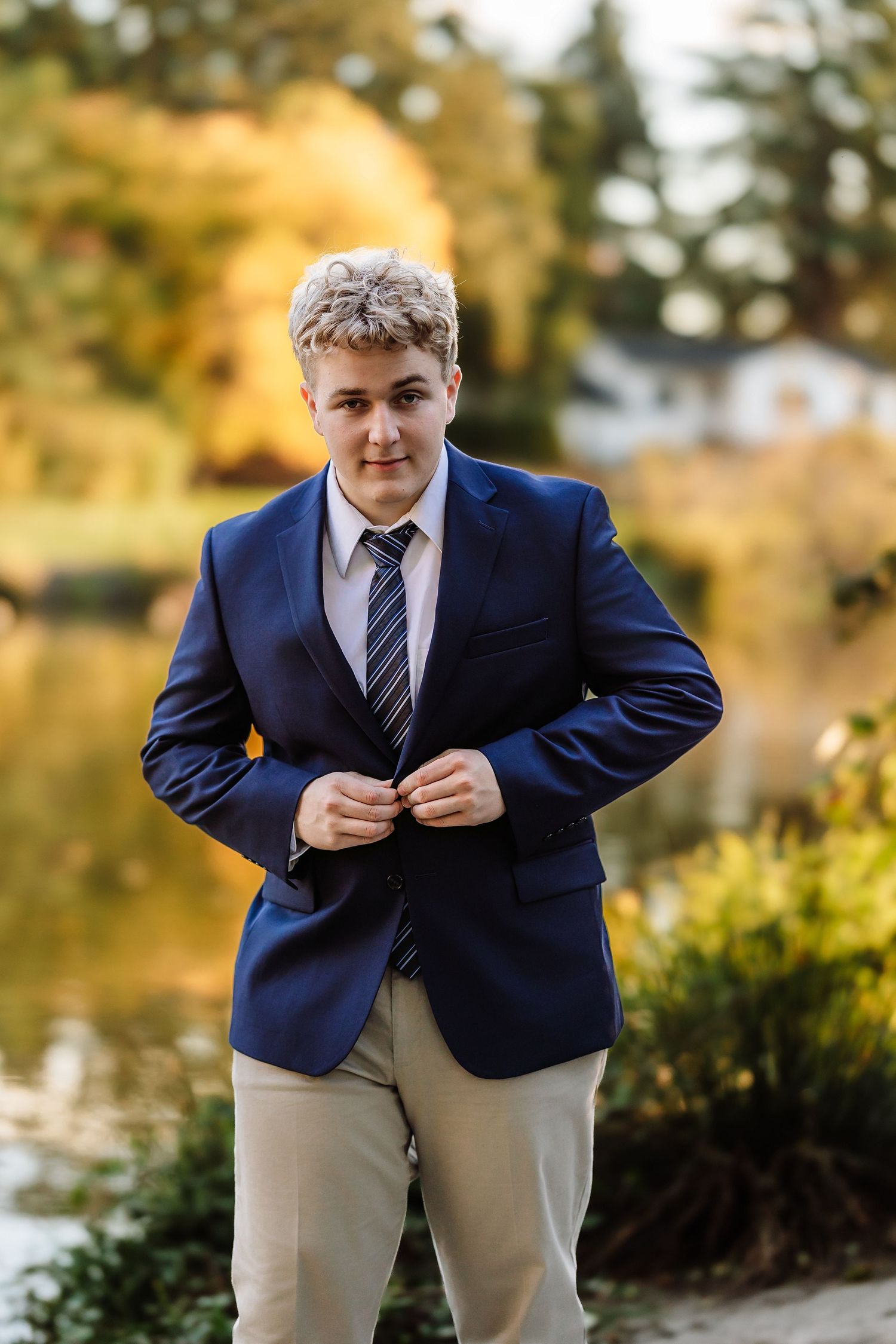Young man in navy blazer and striped tie buttoning jacket outdoors near pond with autumn foliage background.