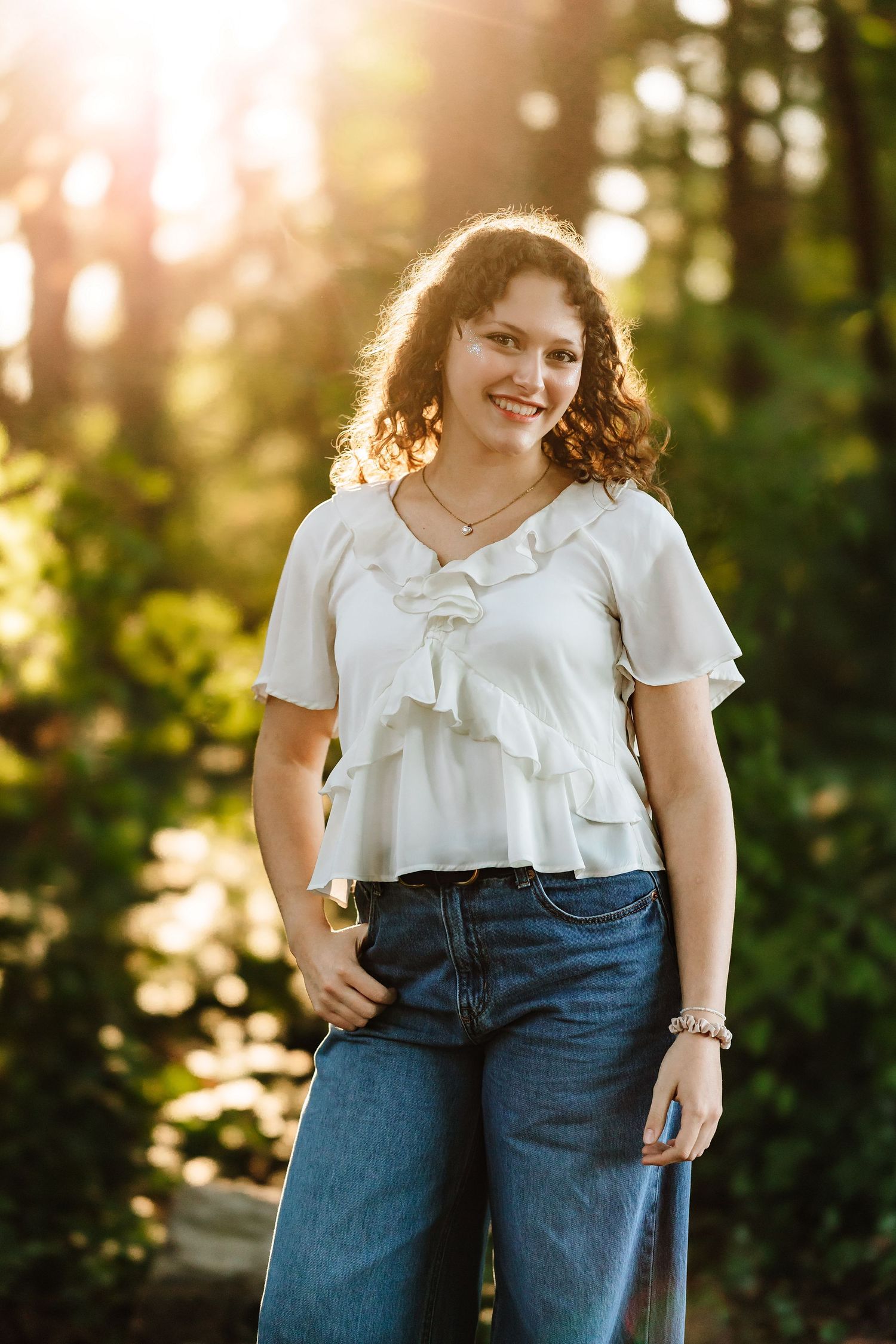 Young woman with curly hair smiles outdoors in white ruffle top and jeans, bathed in golden sunlight.