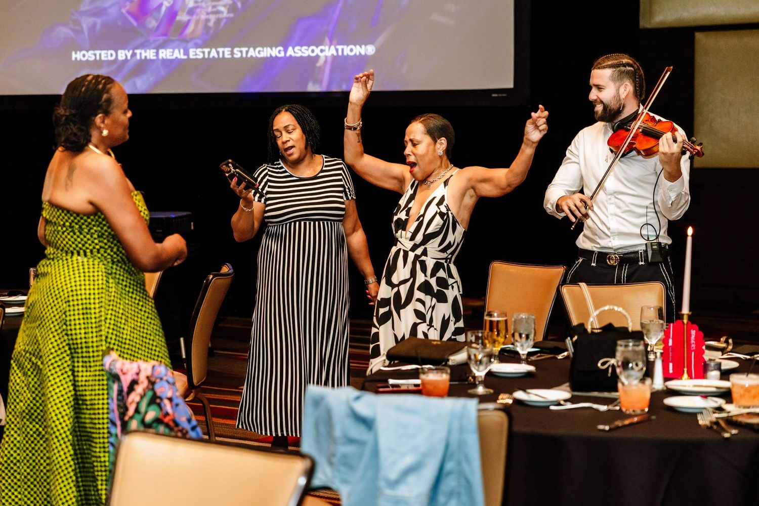 Women dancing at Real Estate Staging Association event with violinist performing live music at elegant banquet.
