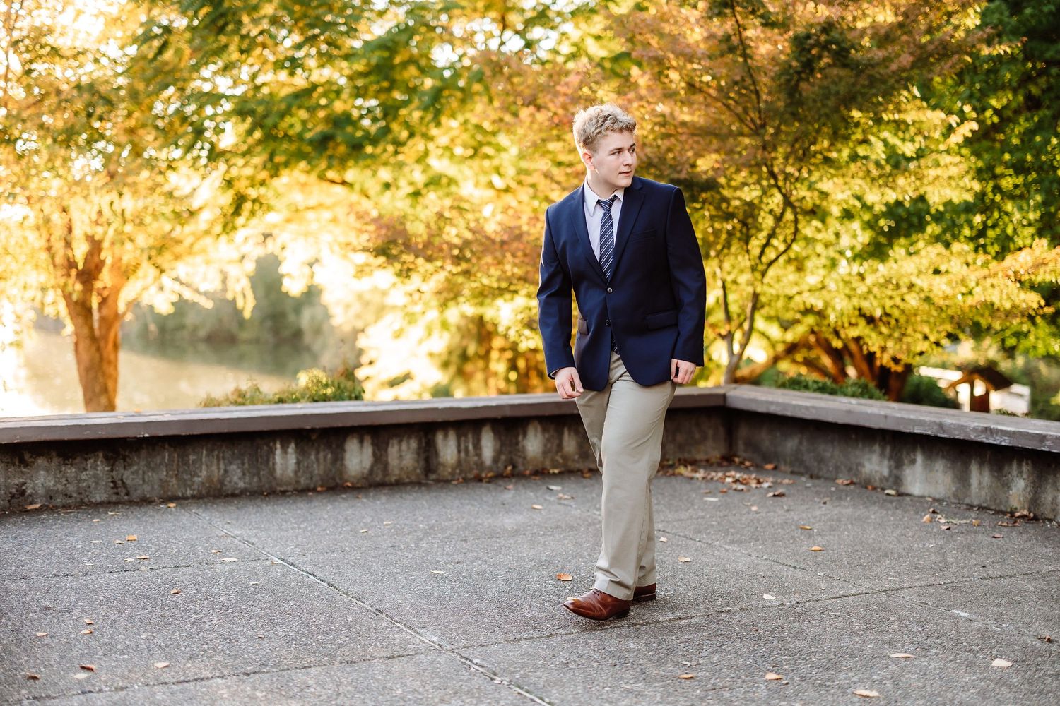 Young man in navy blazer and khaki pants poses outdoors among autumn trees in a professional portrait.
