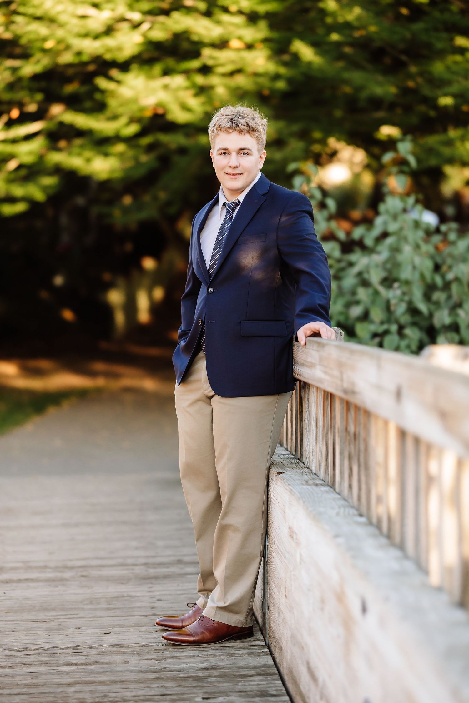 Young man in navy blazer and khakis poses on wooden bridge path surrounded by lush green trees.