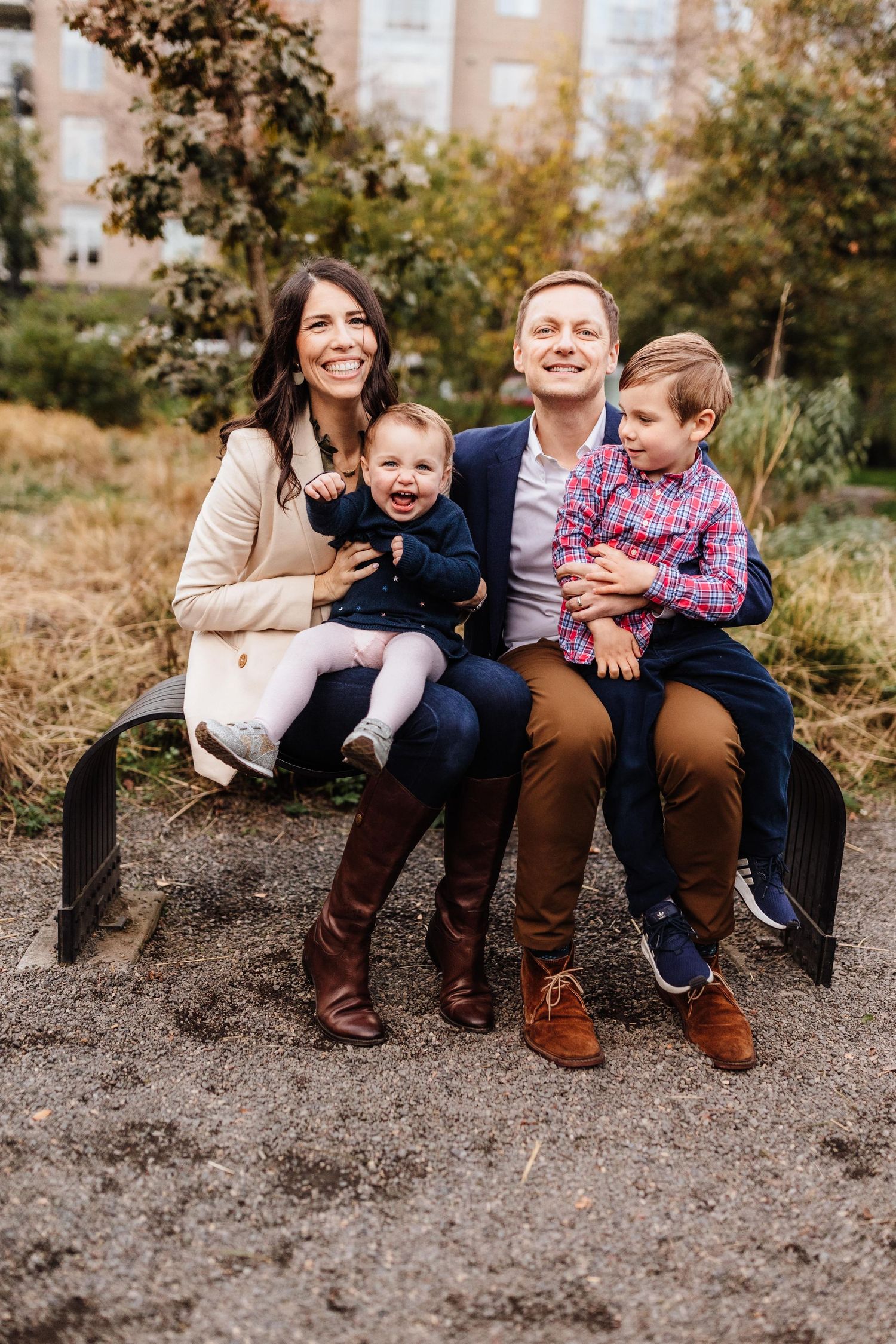 Happy family of four sitting on a bench outdoors in autumn, parents smiling with two young children on their laps.