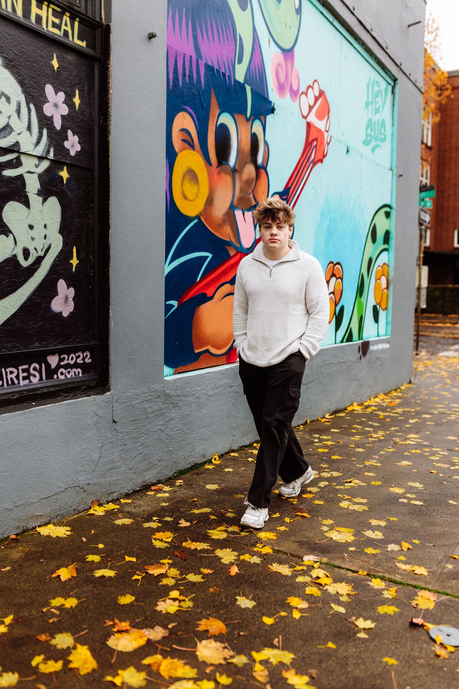 Young man in white hoodie poses on autumn sidewalk covered in fallen leaves beside colorful urban street murals.