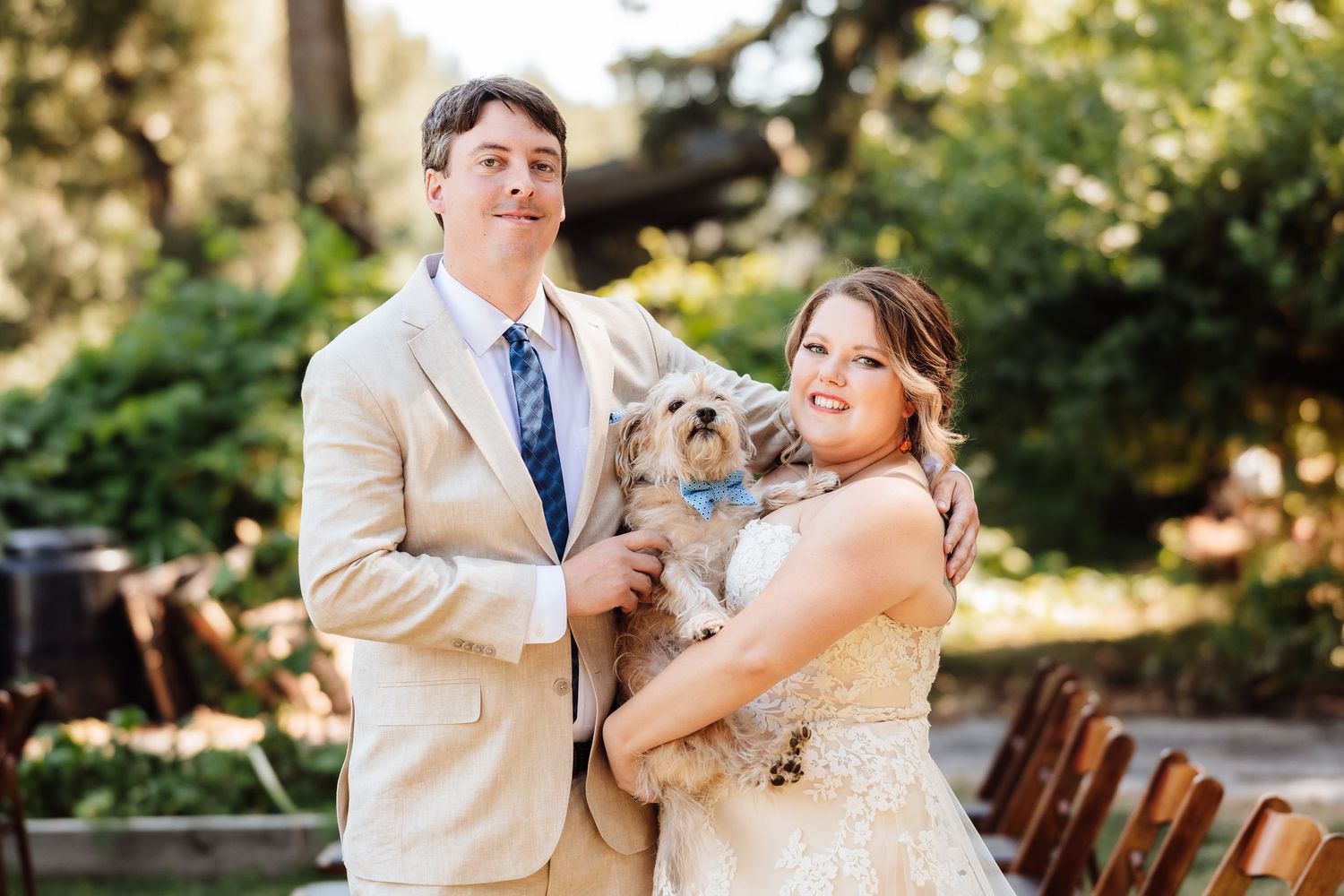 Smiling bride and groom posing with their adorable fluffy dog dressed in a blue bow tie at their outdoor wedding.