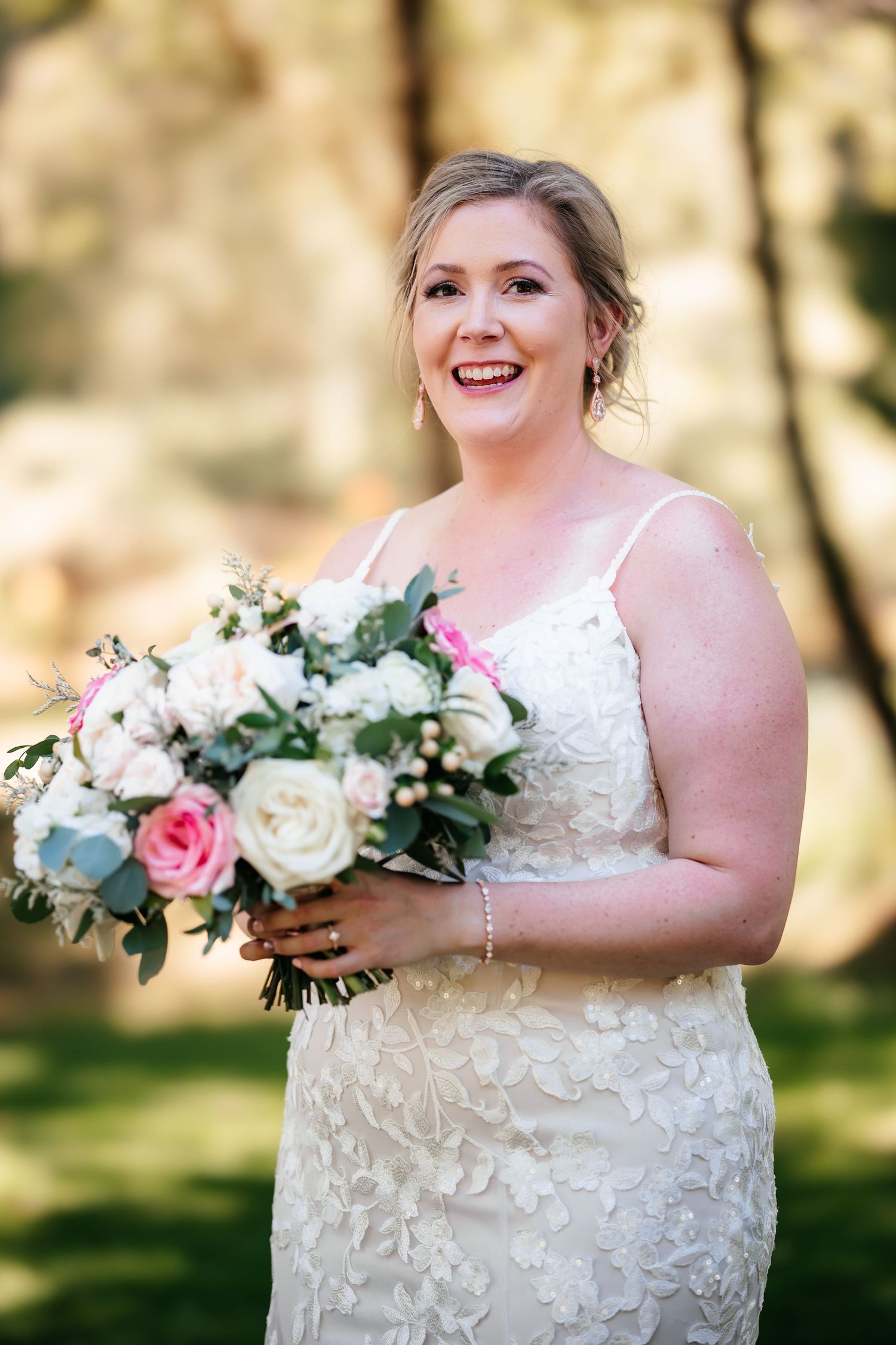 Smiling bride in a lace wedding dress holding a bouquet of pink and white roses outdoors in nature.