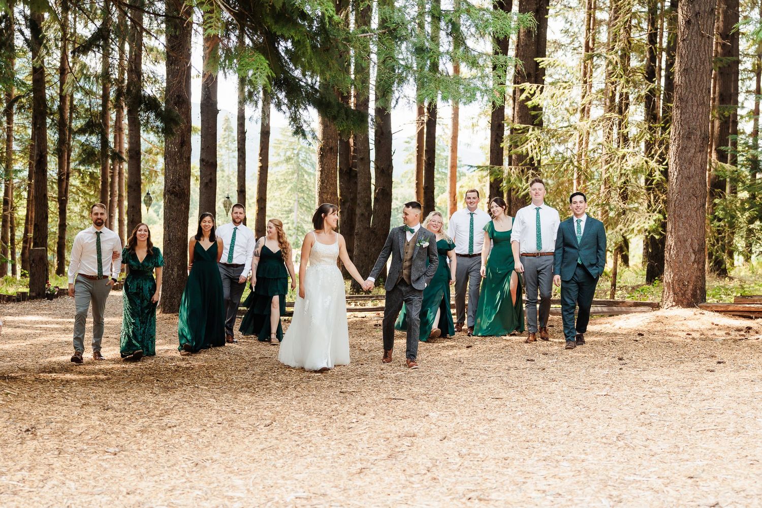 Bride and groom walk with wedding party through sunlit forest, bridesmaids in green dresses, groomsmen in gray.