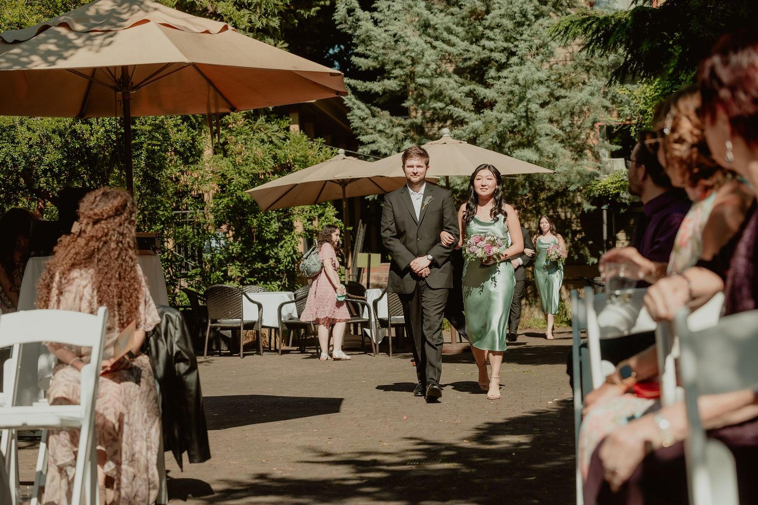 Groomsman escorts bridesmaid in sage dress down sunlit outdoor wedding ceremony aisle.